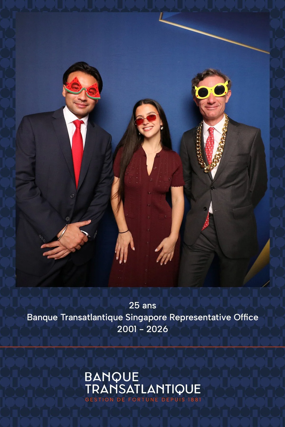 Three people in formal attire wearing humorous sunglasses at a celebration. The man on the left is wearing watermelon-shaped sunglasses, the woman in the middle is wearing red-tinted sunglasses, and the man on the right is wearing yellow, sunglasses with a chain necklace. The background is blue, and text at the bottom indicates the event is for the Bank Transatlantique Singapore Office's 25th anniversary from 2001 to 2026.
