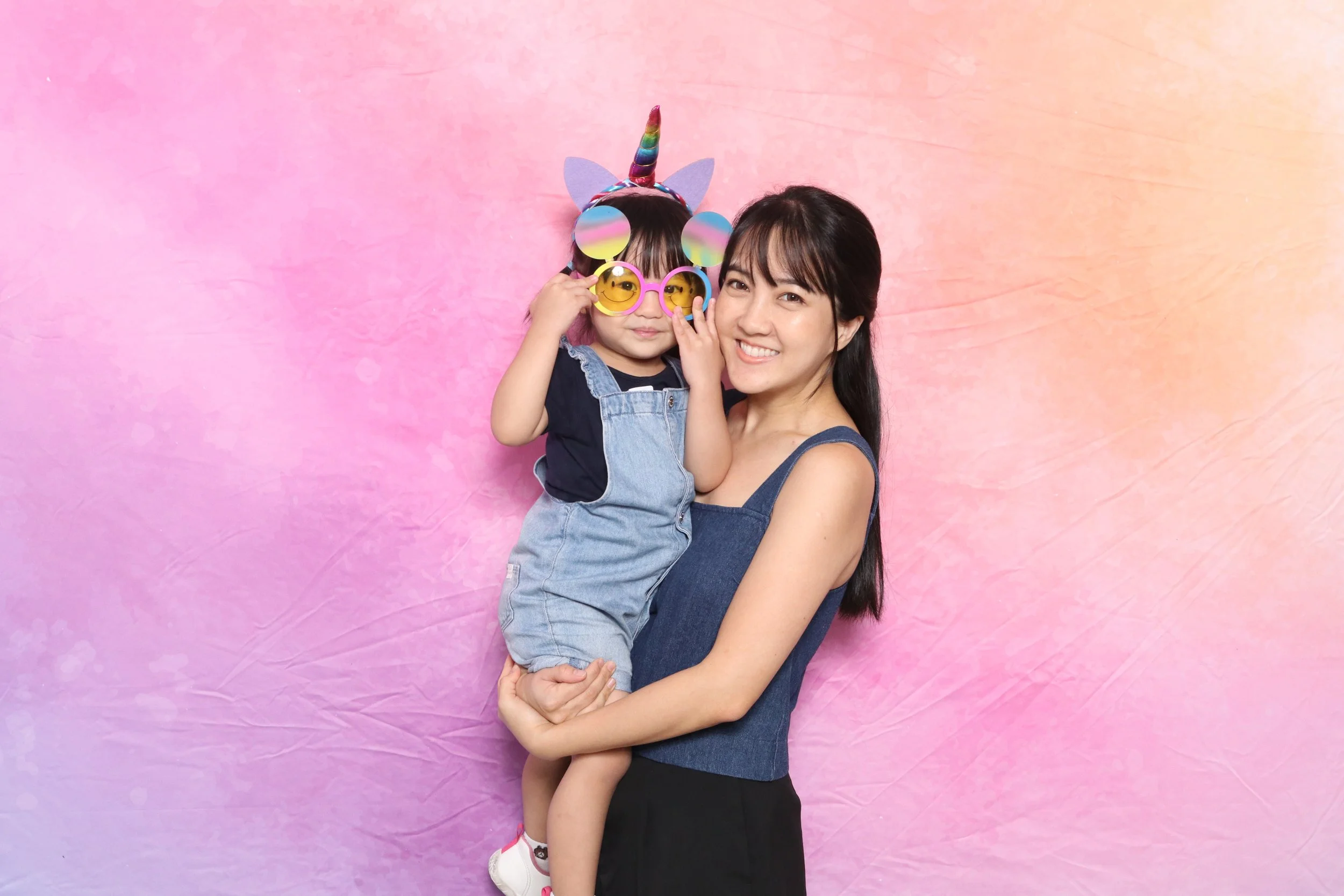 A woman holding a young girl who is wearing rainbow unicorn and smiley face sunglasses, with a pink and peach watercolor background.
