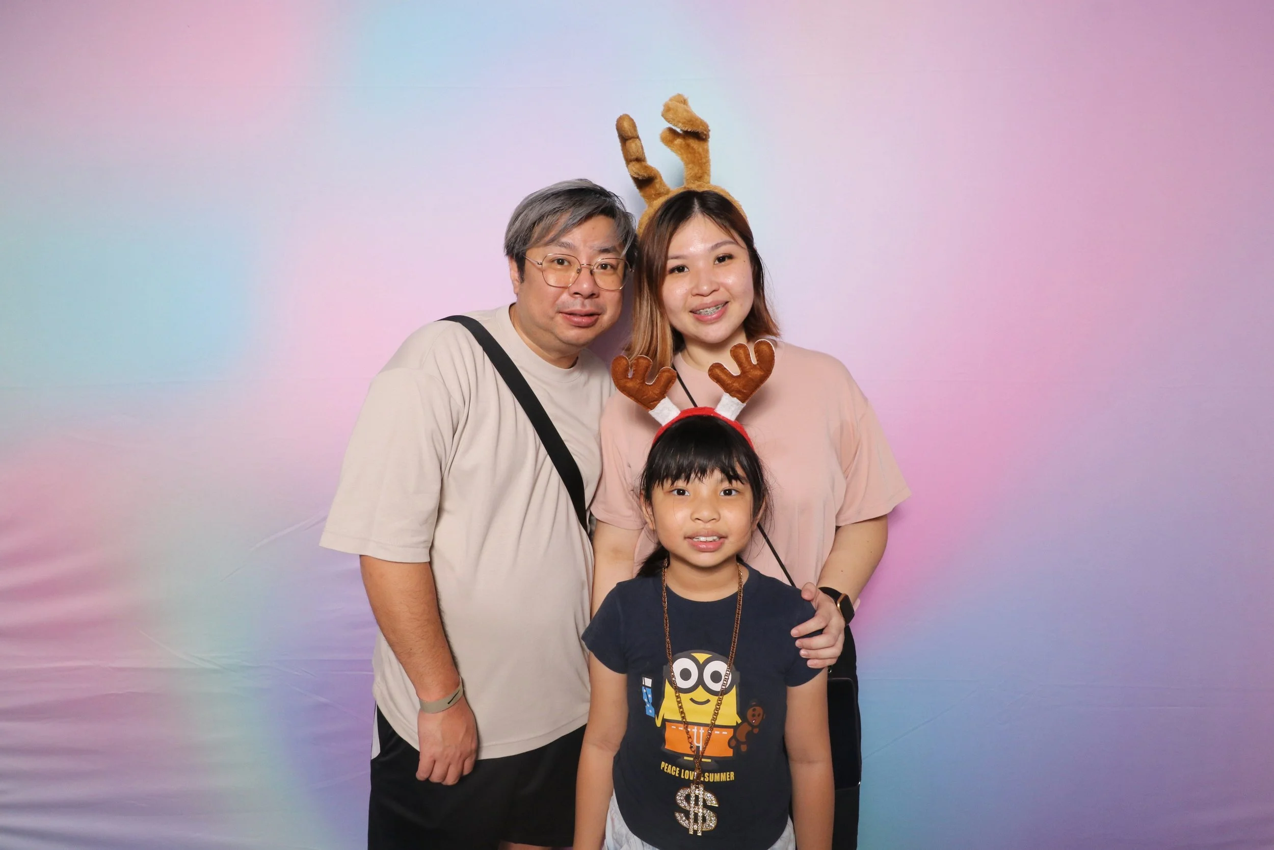 Family of three posing in front of a pastel rainbow background, with the mother and father wearing reindeer headbands and the daughter wearing a Minions t-shirt with a gold chain necklace with a dollar sign pendant.
