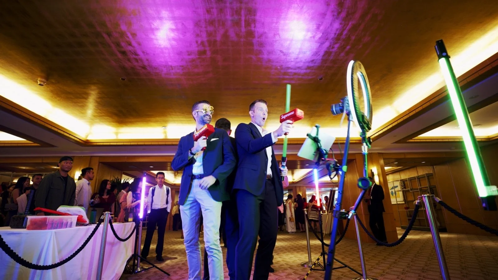 Two men in suits holding toy laser guns with the Supreme logo, posing playfully at a party or event with a crowd and colorful lighting in the background.