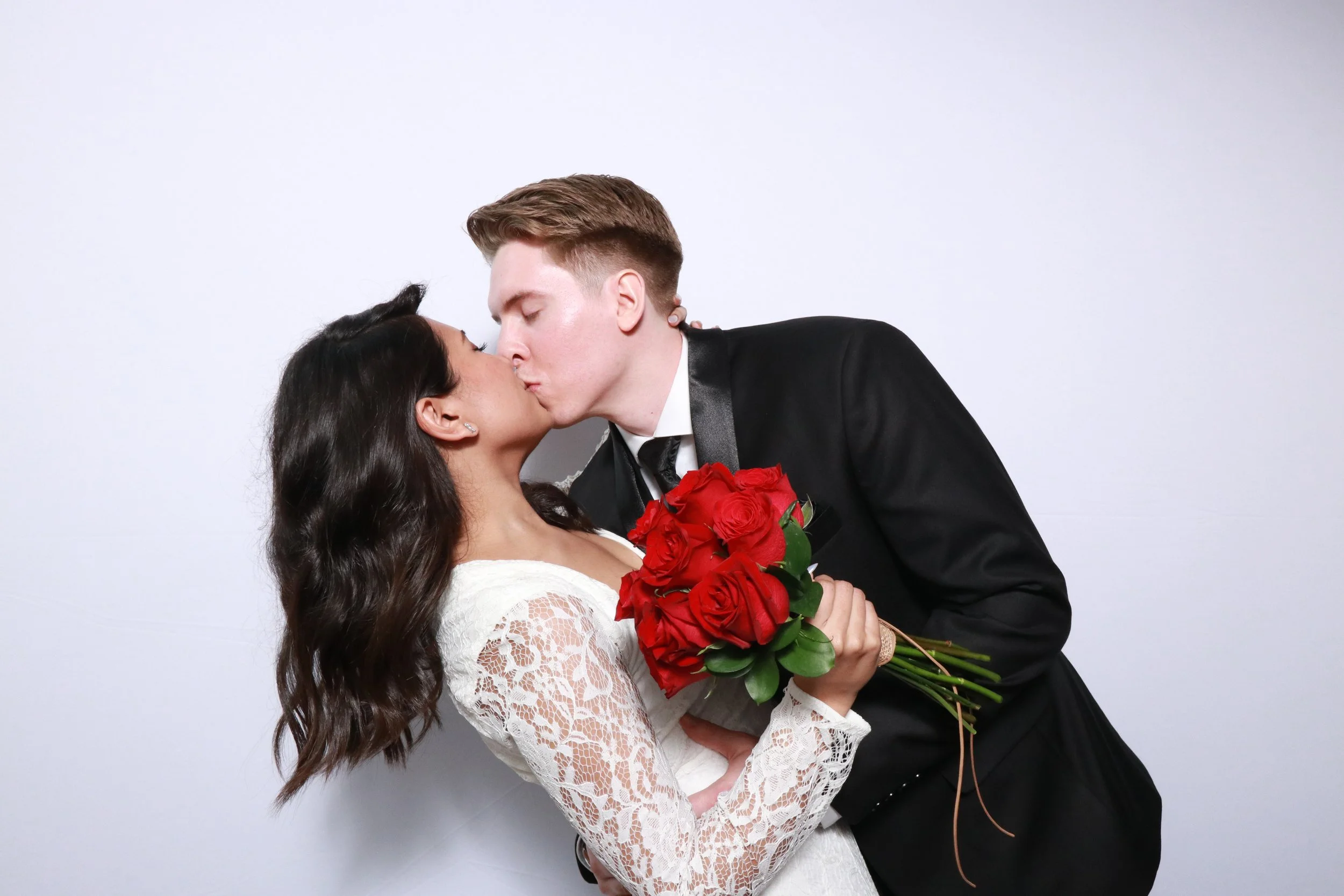 A couple kissing at their wedding, with the bride holding a bouquet of red roses, and the groom dressed in a black tuxedo with a red boutonniere, against a plain white background.