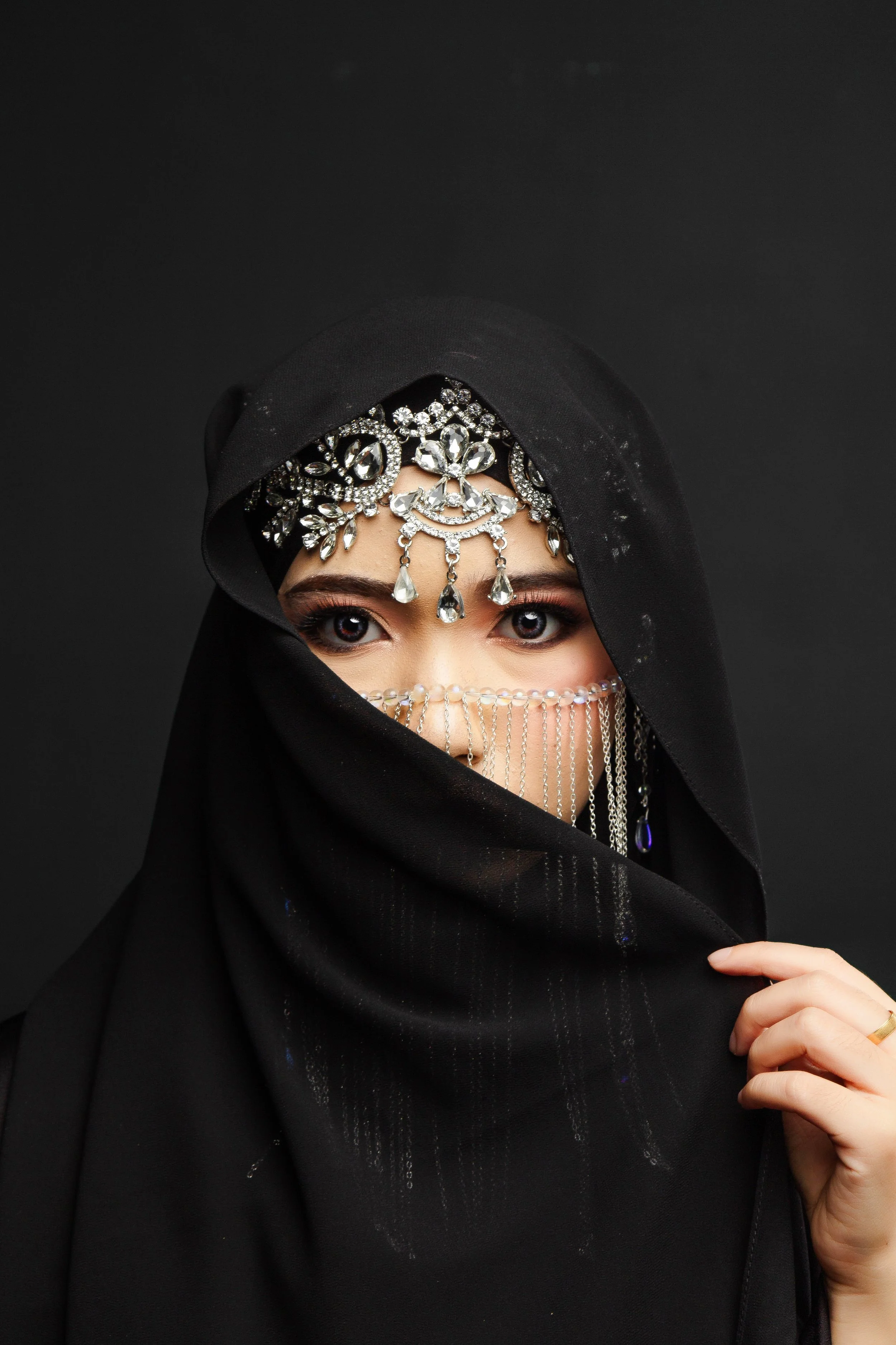 A woman wearing a black head covering and black dress, adorned with large, ornate silver jewelry with crystals on her forehead, around her head, and hanging down her face, against a black background.