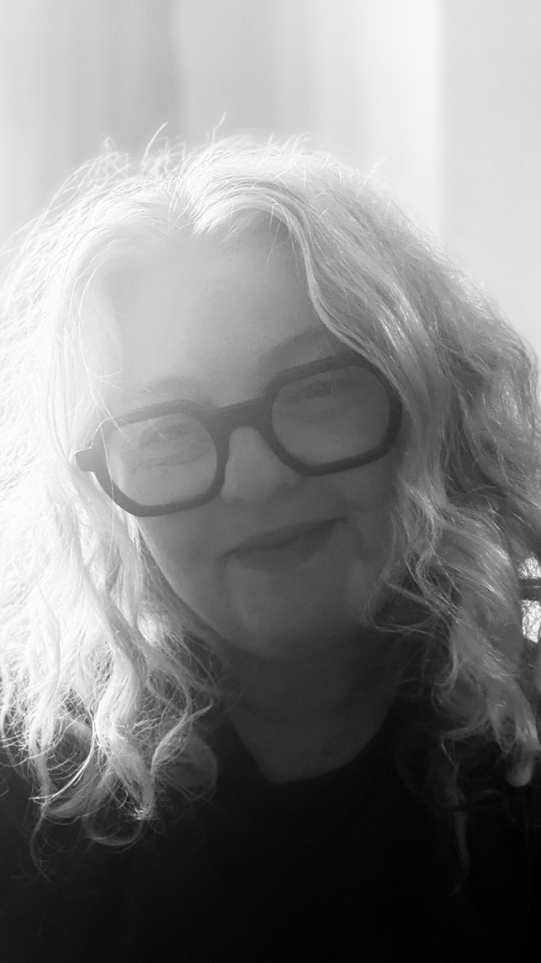 A woman with long, curly hair wearing large, dark glasses, smiling in a black-and-white photo.