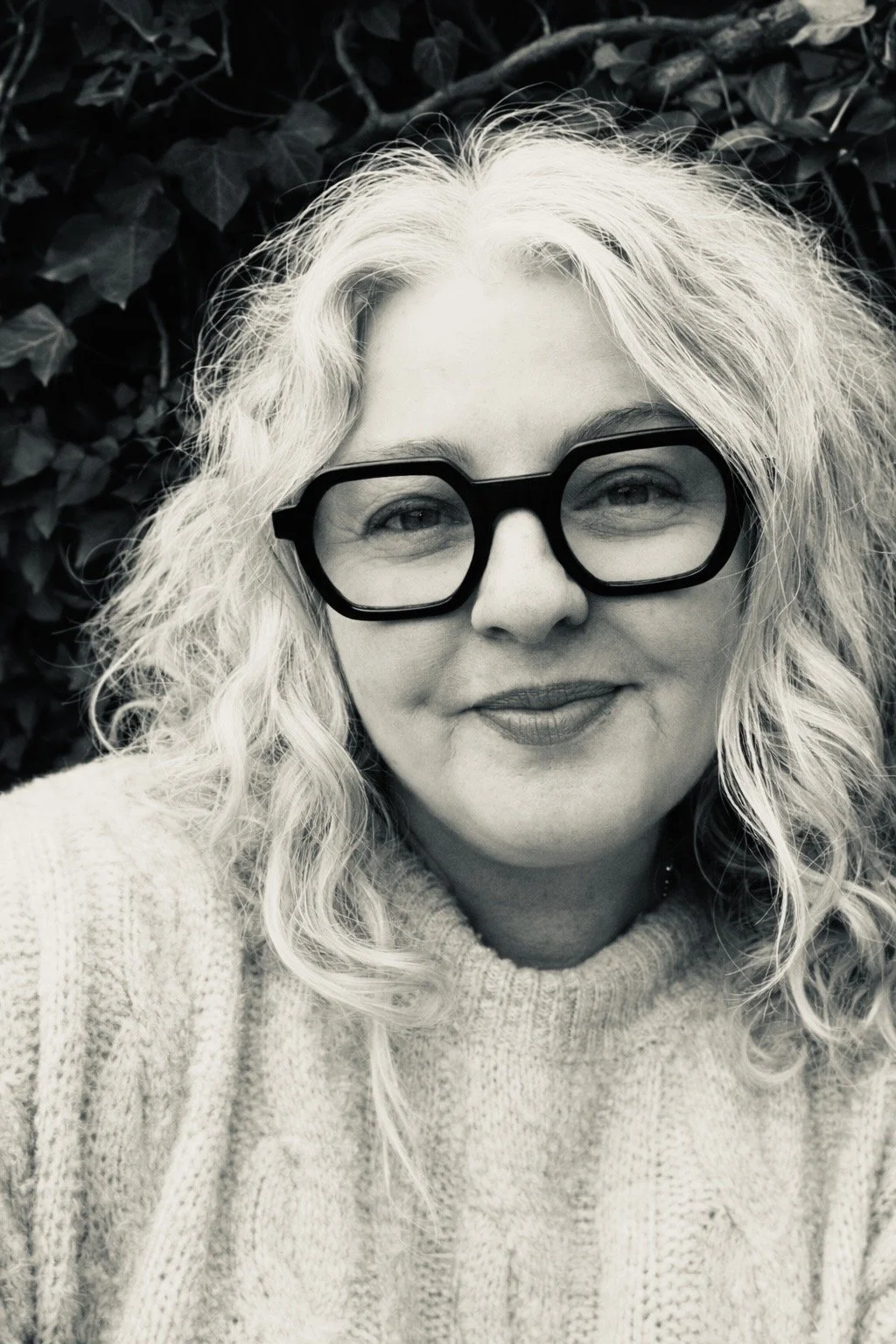 A woman with curly blonde hair and large black glasses smiling at the camera, wearing a cozy knit sweater, with dark foliage in the background.