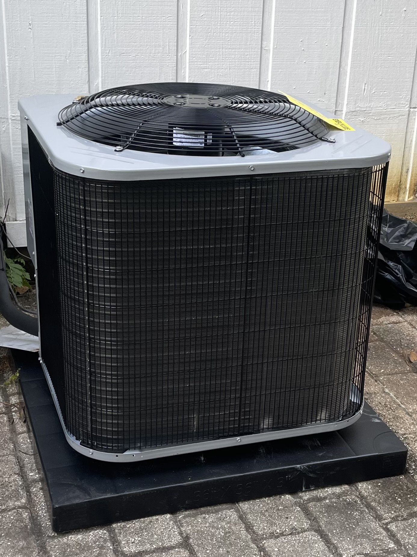 An outdoor air conditioning unit with a large fan on top, surrounded by a black protective grille, placed on a concrete surface against a white wooden wall.
