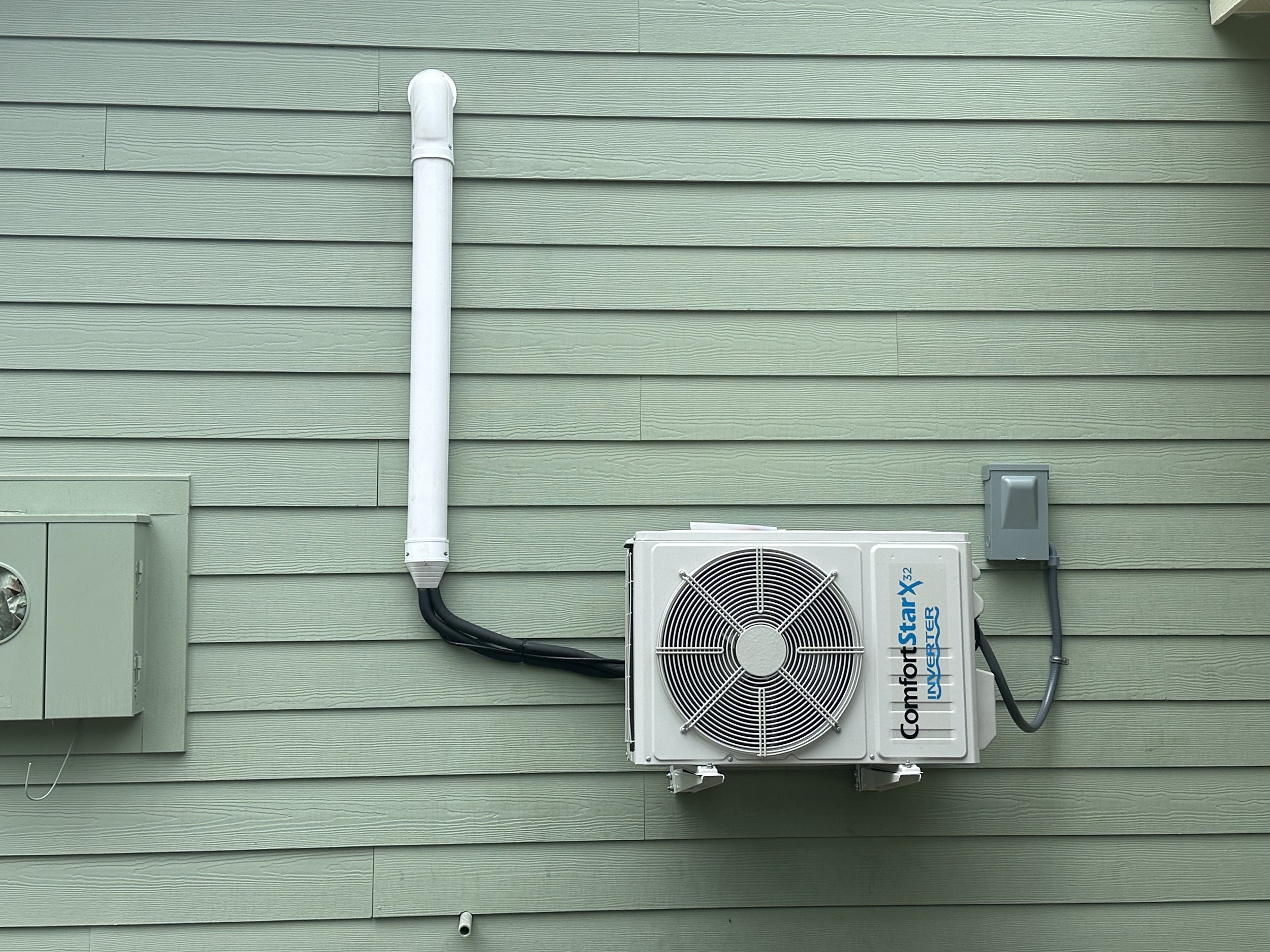 Exterior of a house with light green vinyl siding, featuring a white drainpipe running vertically down the wall and an air conditioning unit mounted on the wall with black and white pipes connected.