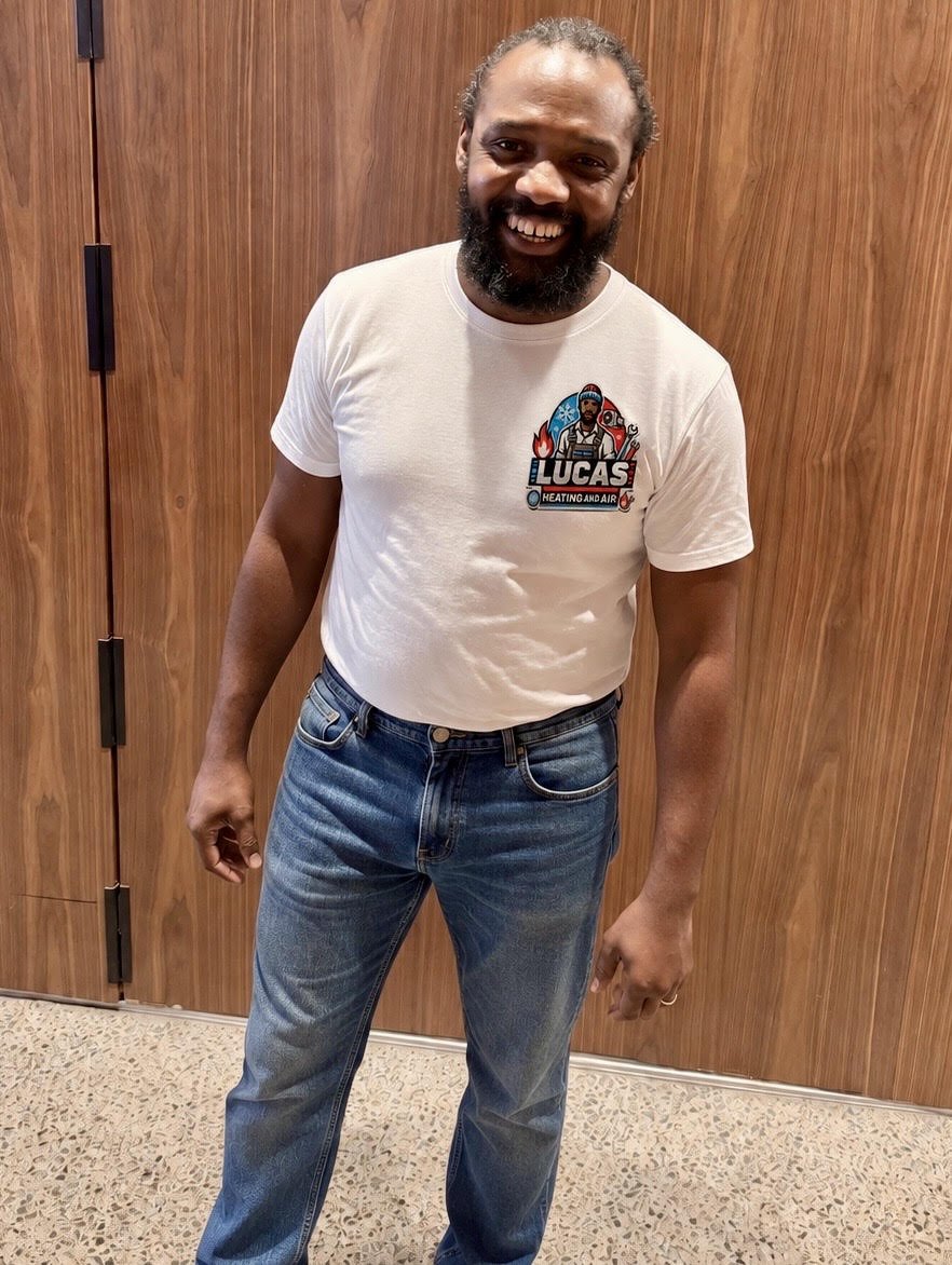 Man with a beard smiling, wearing a white t-shirt with a logo that says "LUCAS Heating and Air" and jeans, standing in front of a wooden wall.