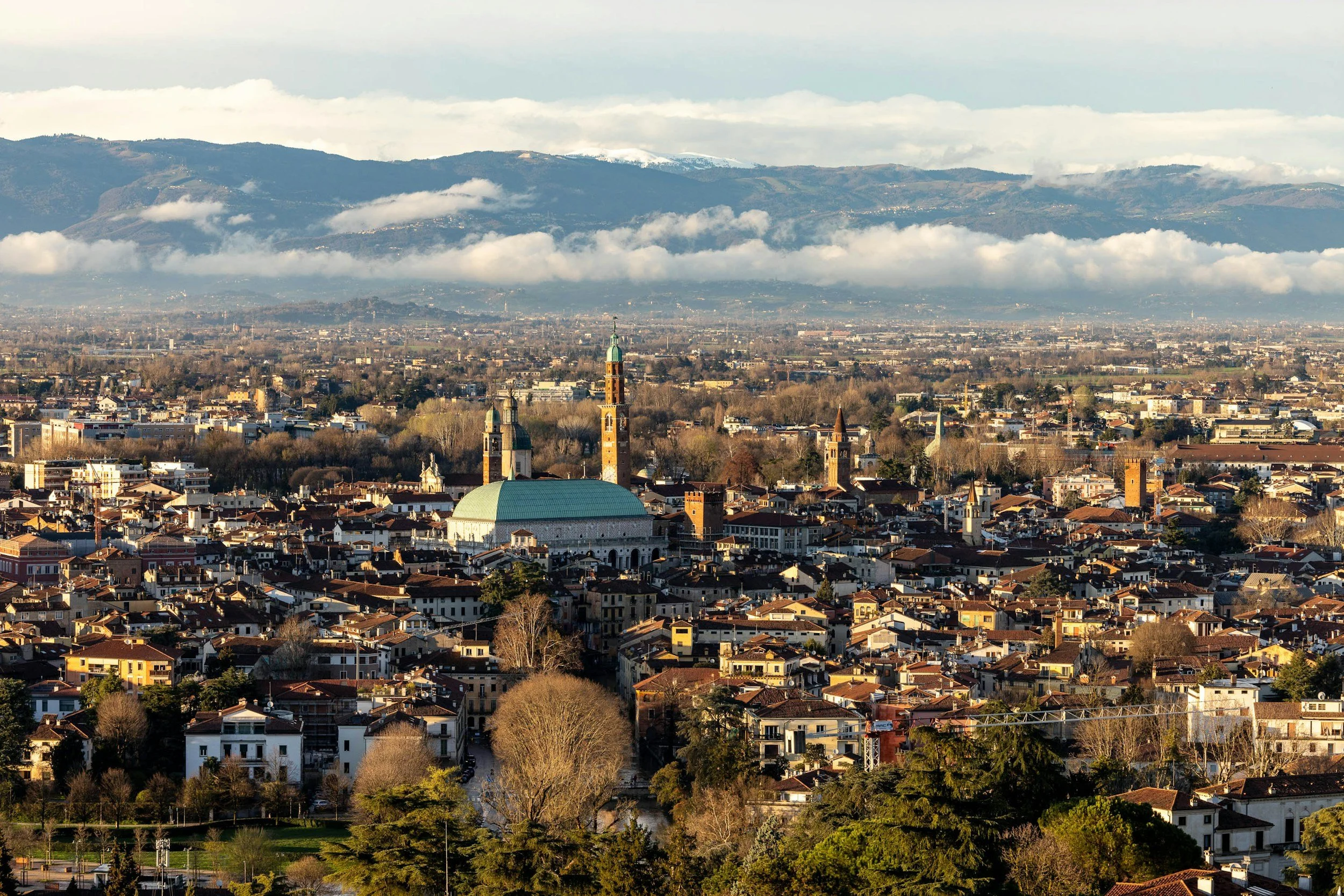Paesaggio urbano con chiese, case e alberi sullo sfondo di montagne con alcune cime innevate e nuvole.