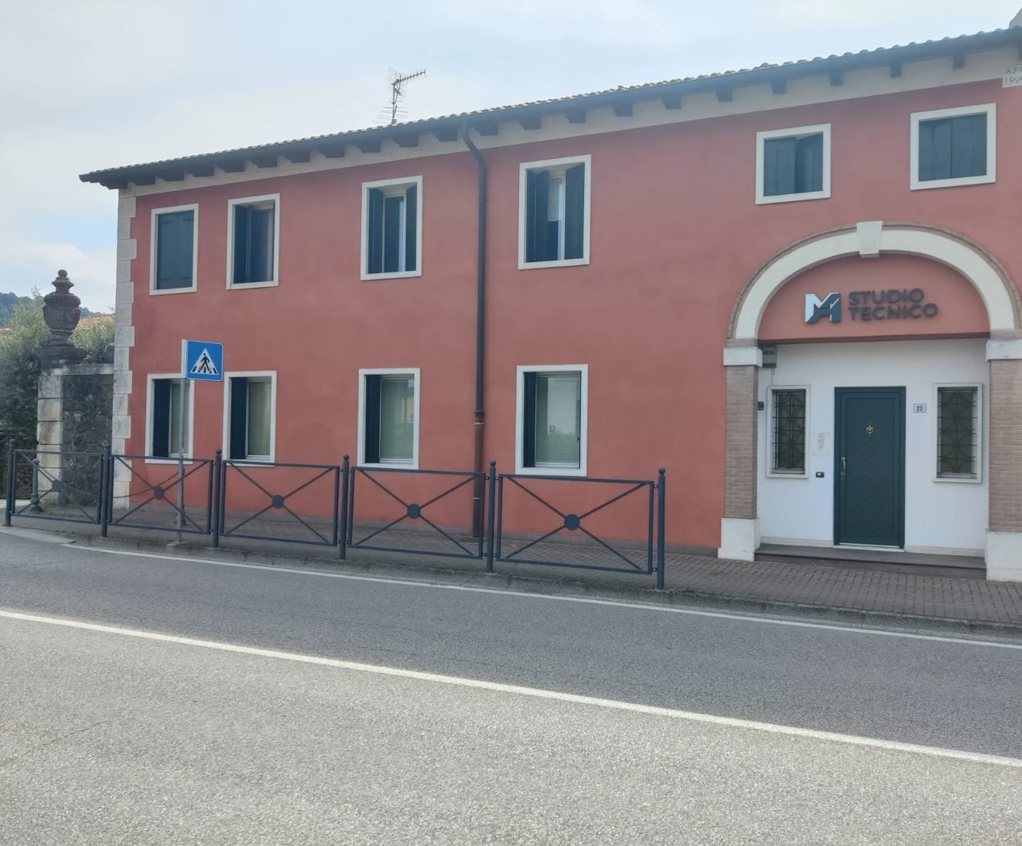 Edificio color pesca con segnale di attraversamento pedonale, facciata con finestra, ingresso principale con scritta 'Studio Tecnico Muraro' sopra, ufficio numero 23, strada e marciapiede in primo piano.