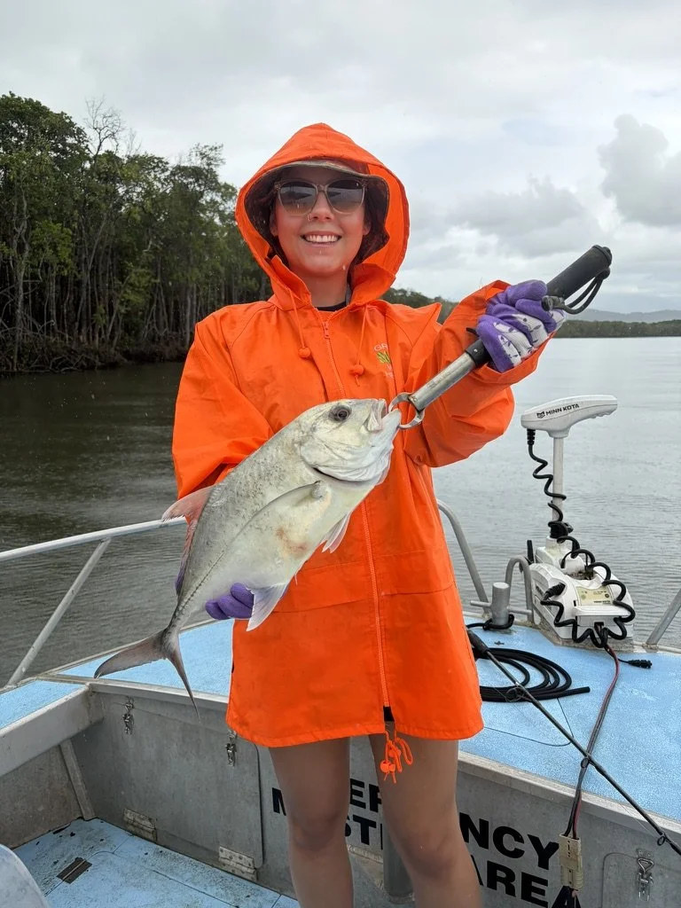 Woman in orange raincoat and sunglasses holding a large fish on a boat with water and trees in the background.