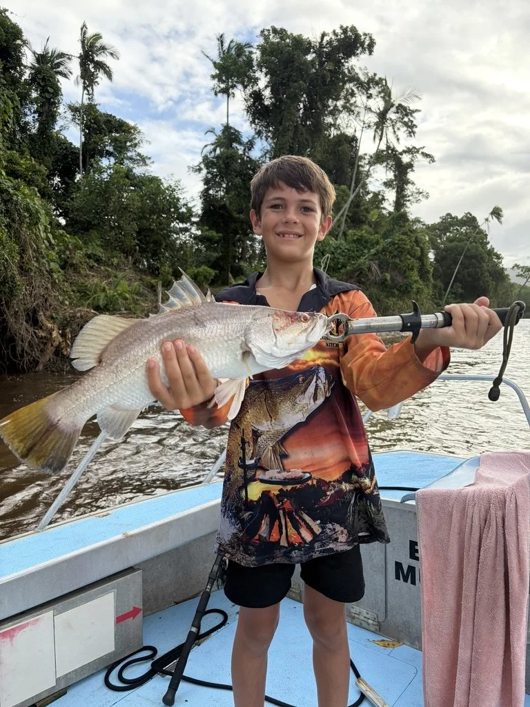 A boy on a boat holding a large fish with a fishing rod in the background, with trees and cloudy sky behind him.
