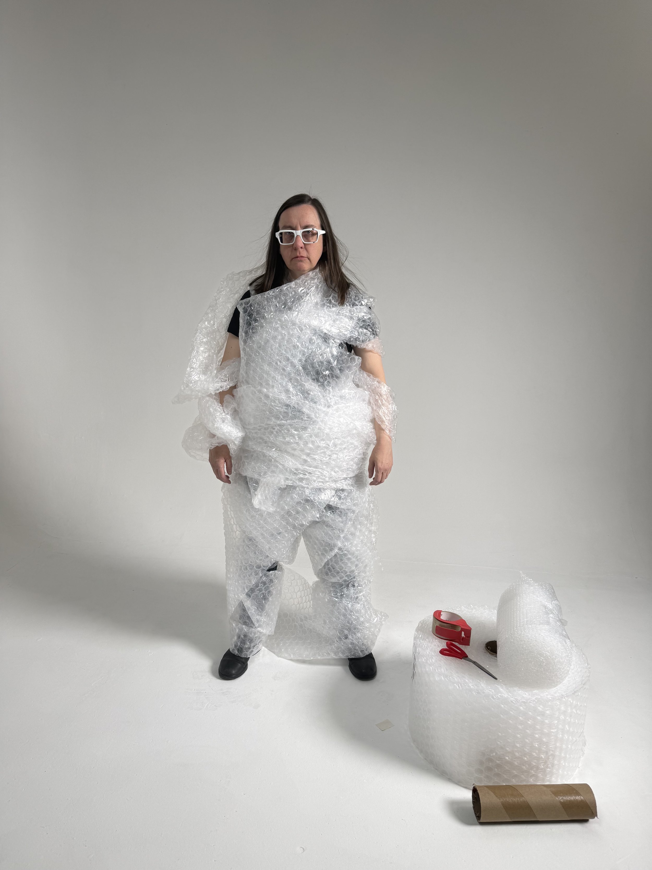 A woman wearing glasses and black clothing standing in front of a plain white background, wrapped in and surrounded by clear bubble wrap and packing materials, with scissors and packing tape on the floor nearby.