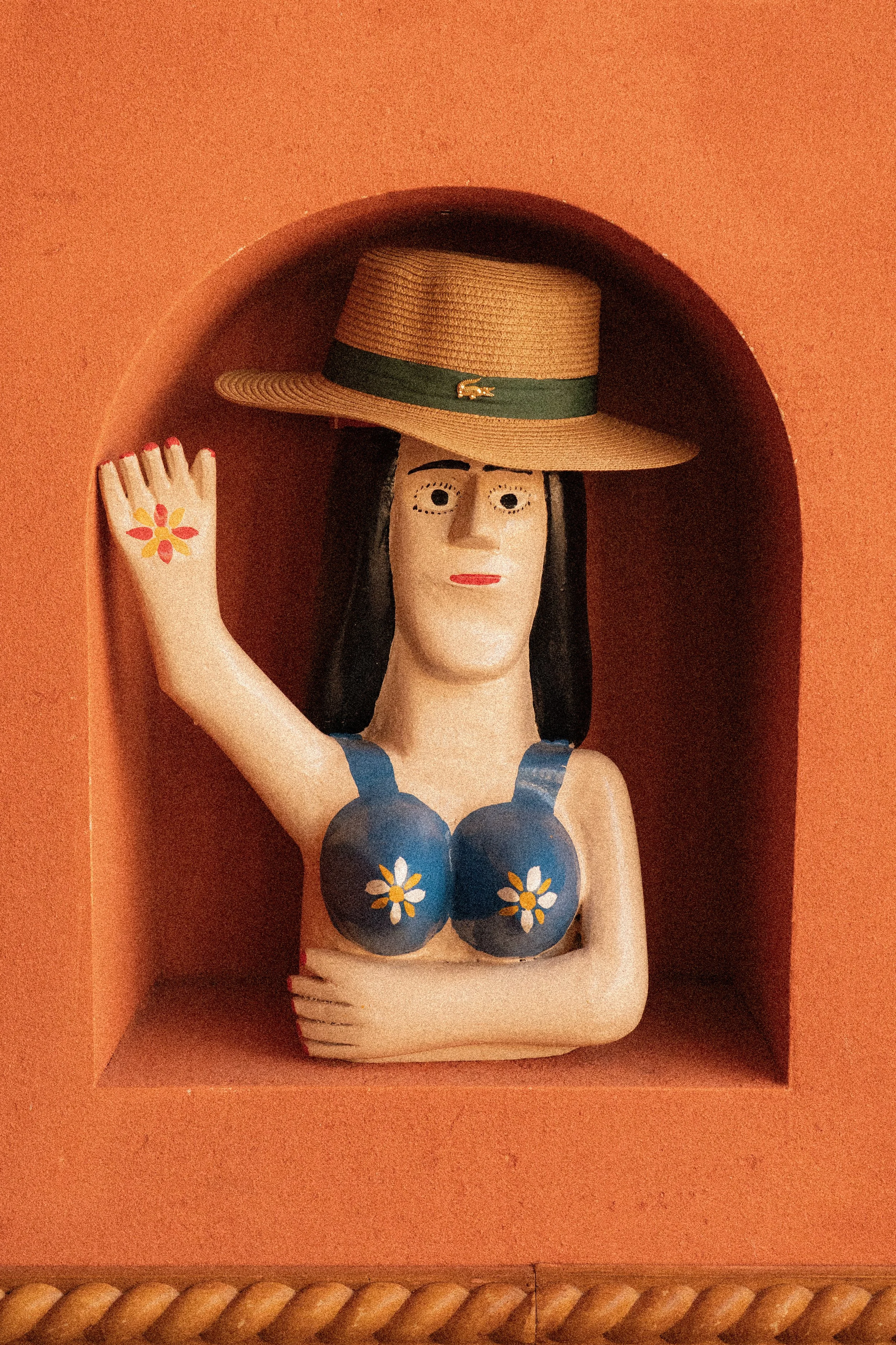 A wooden sculpture of a woman with long black hair, wearing a blue top with white and yellow flowers, a wide-brimmed straw hat with a green band and a small gold detail, and red nails. The sculpture is inside a pinkish-orange display case with a curved top and a rope border at the bottom.