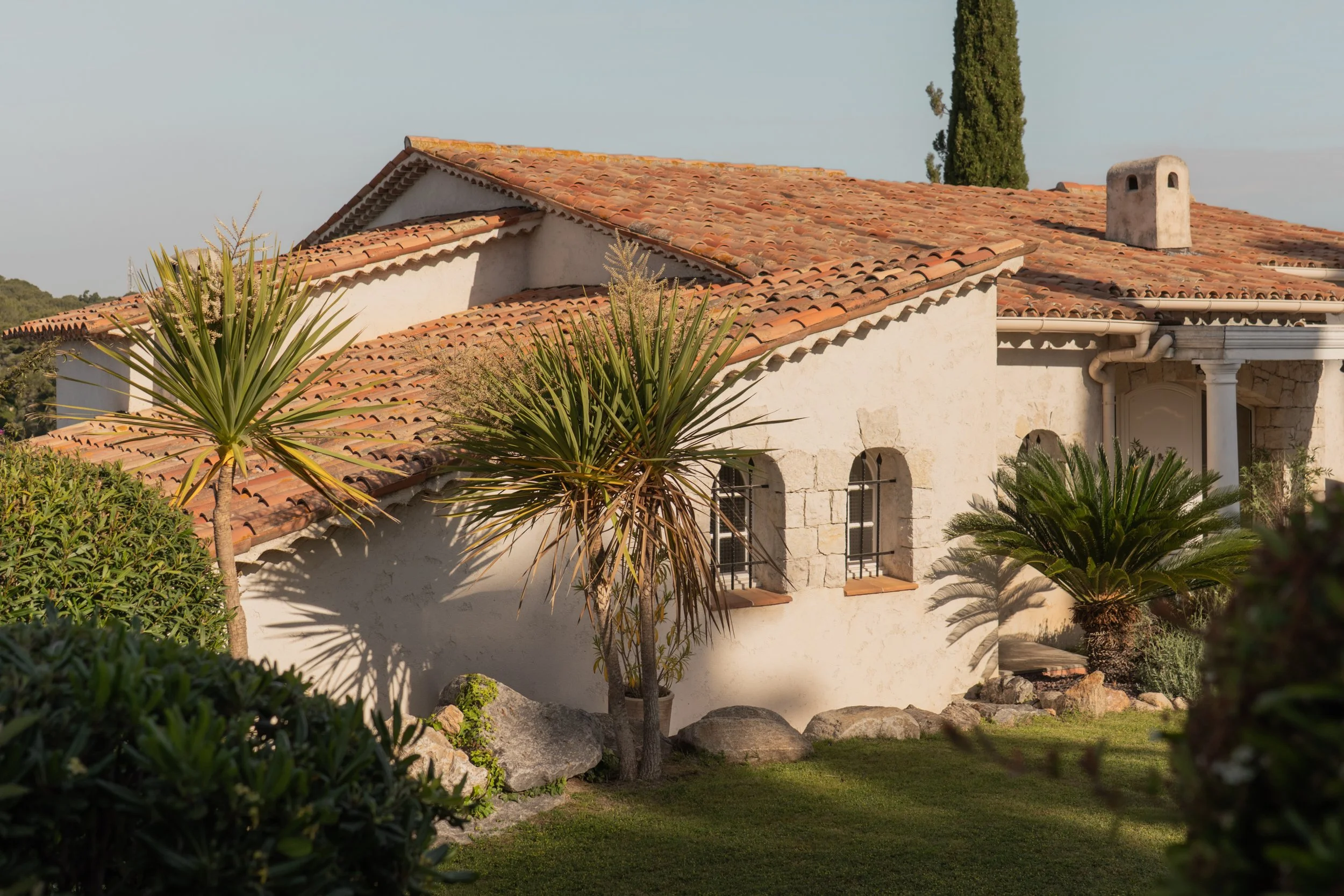 A Mediterranean-style house with a red-tiled roof, surrounded by lush greenery and tall trees, with rocks and a well-maintained lawn in the foreground.
