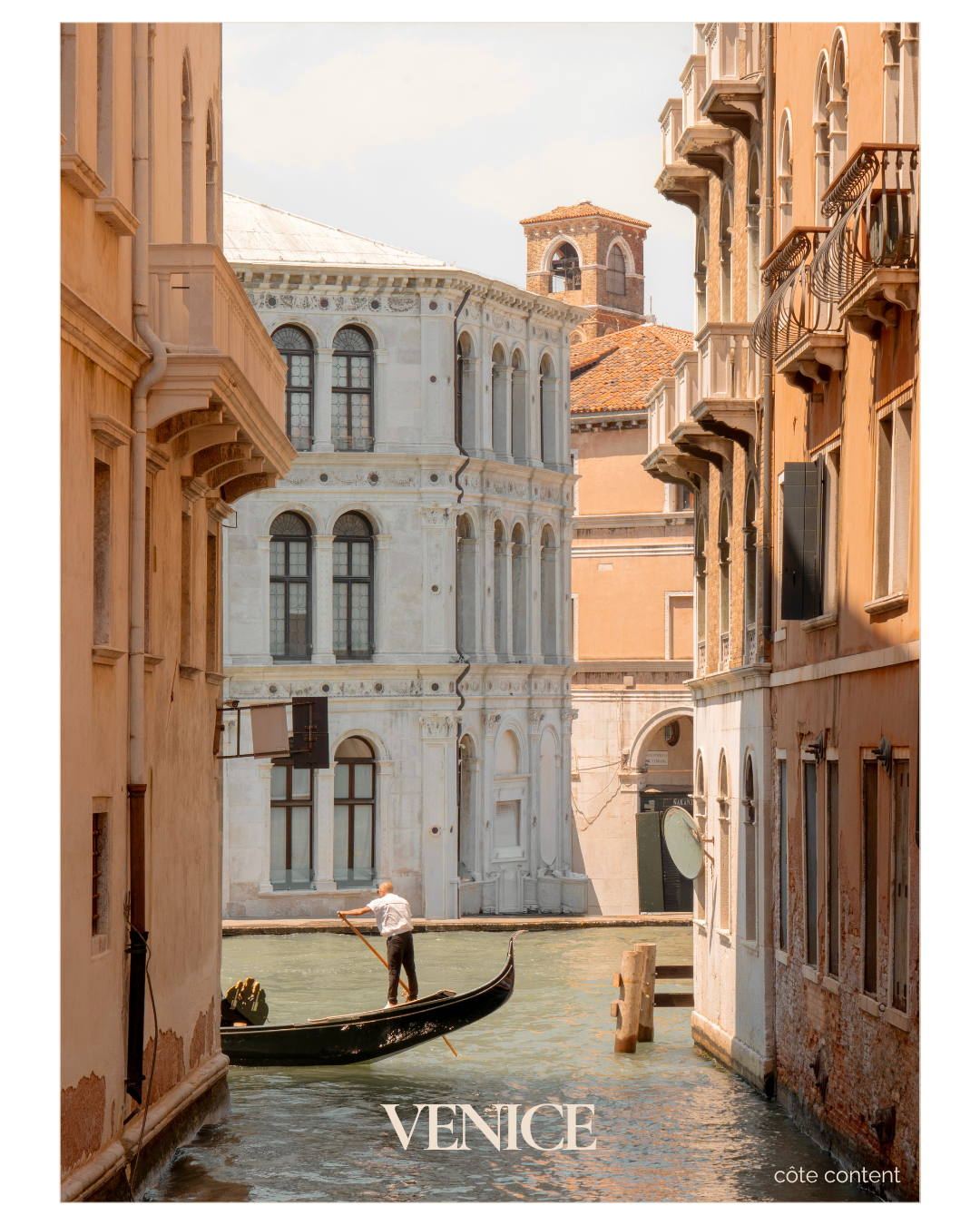 A gondolier in a white shirt and dark pants standing in a black gondola on a canal in Venice, Italy, surrounded by historic buildings with warm-colored facades and detailed architecture.