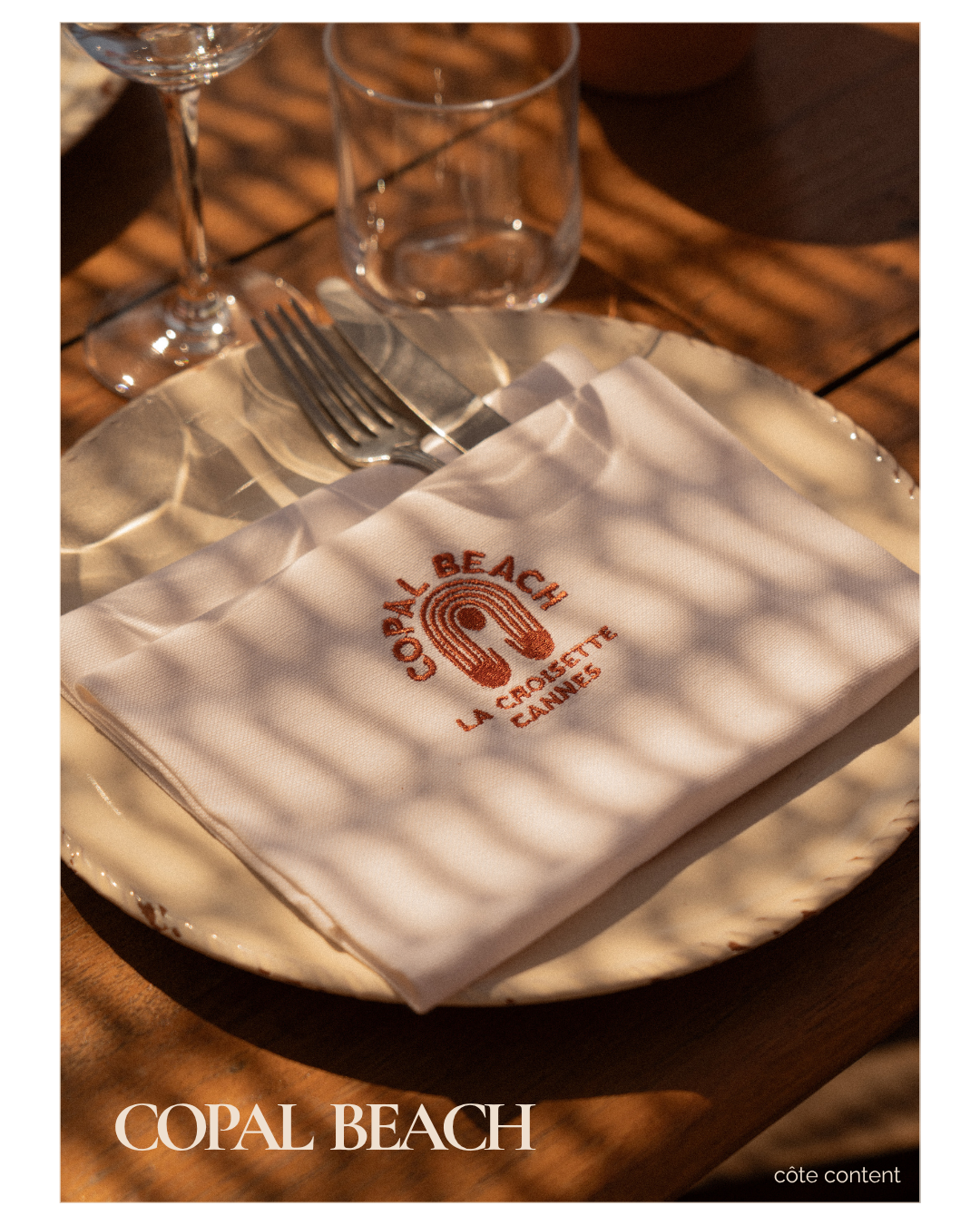 A place setting on a wooden table with a cream-colored plate, a folded white napkin embroidered with 'Copal Beach La Croisette Cannes,' a fork resting on the napkin, and two glasses in the background, with shadows cast by nearby foliage.