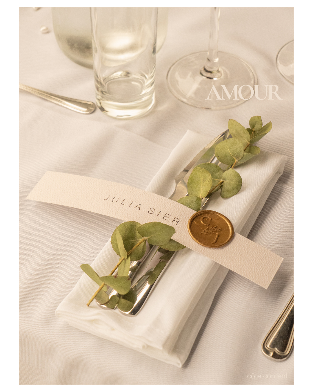 Elegant table setting featuring a wrapped napkin with a sprig of eucalyptus, a name card with 'Julia Sier,' a gold wax seal with initials, and glassware on a white tablecloth.