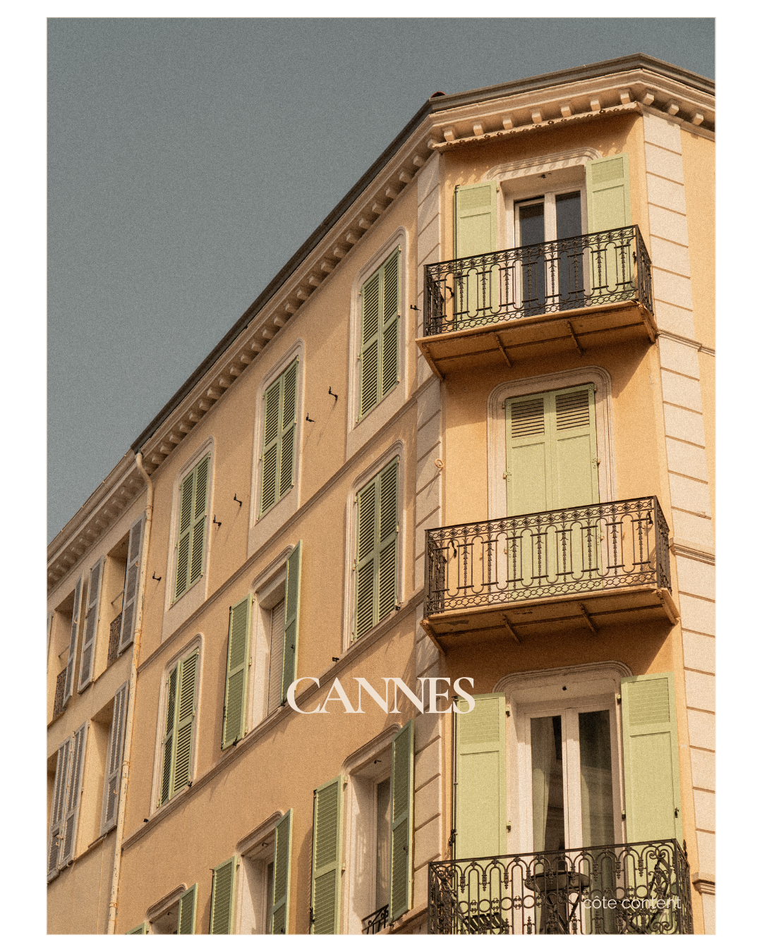 A close-up view of a multi-story building in Cannes with beige walls, green window shutters, and decorative wrought-iron balcony railings, under a clear sky.