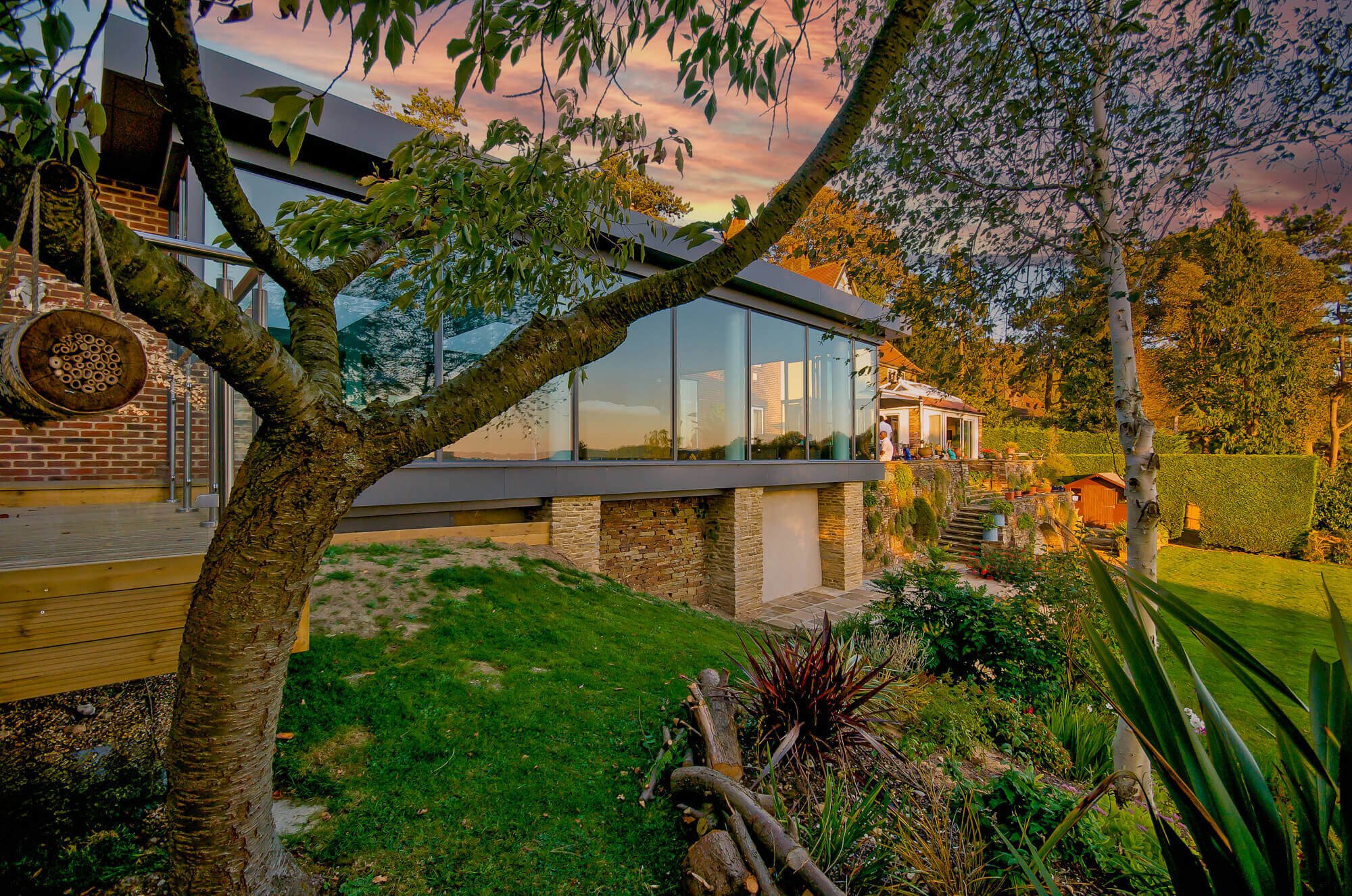 A modern house with large glass windows and a backyard garden during sunset, featuring trees, plants, and outdoor furniture.