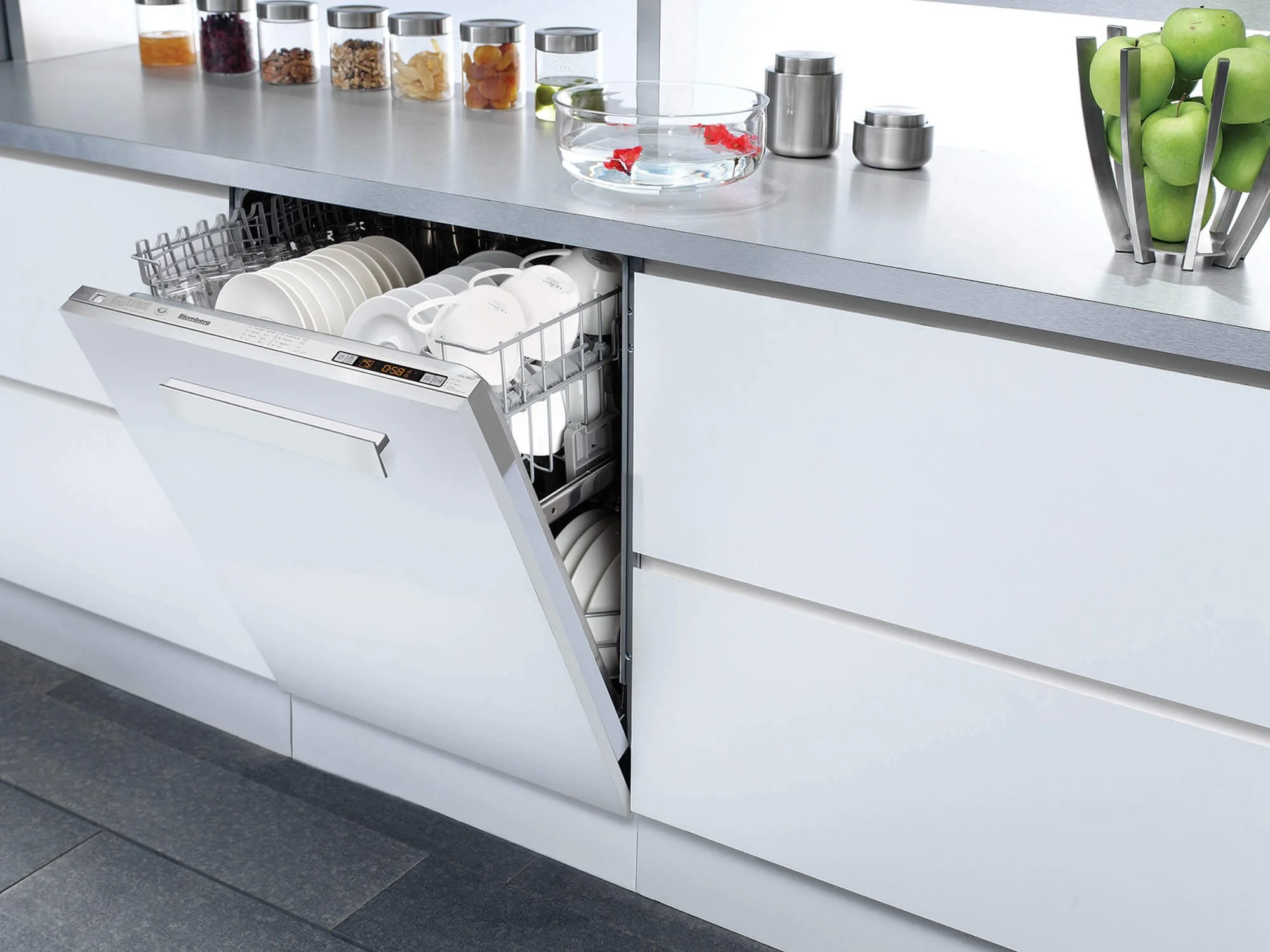 Open dishwasher filled with clean white dishes, mugs, and bowls in a modern kitchen with a gray countertop, jars of dried goods, and a metal fruit holder with green apples.