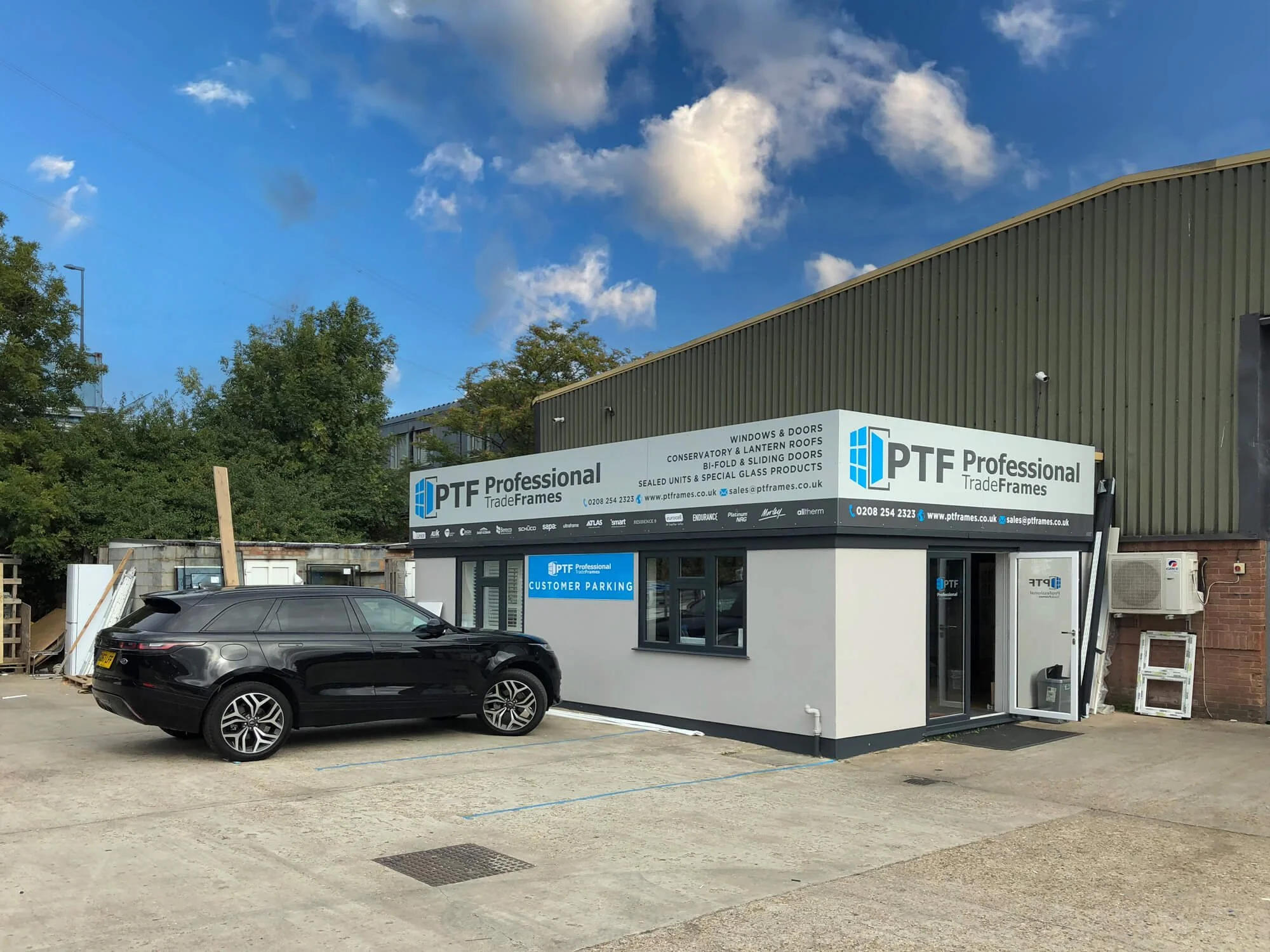 Outside view of PTF Professional Trade Frames building with a black car parked in front, clear blue sky with some clouds, and parking lot.