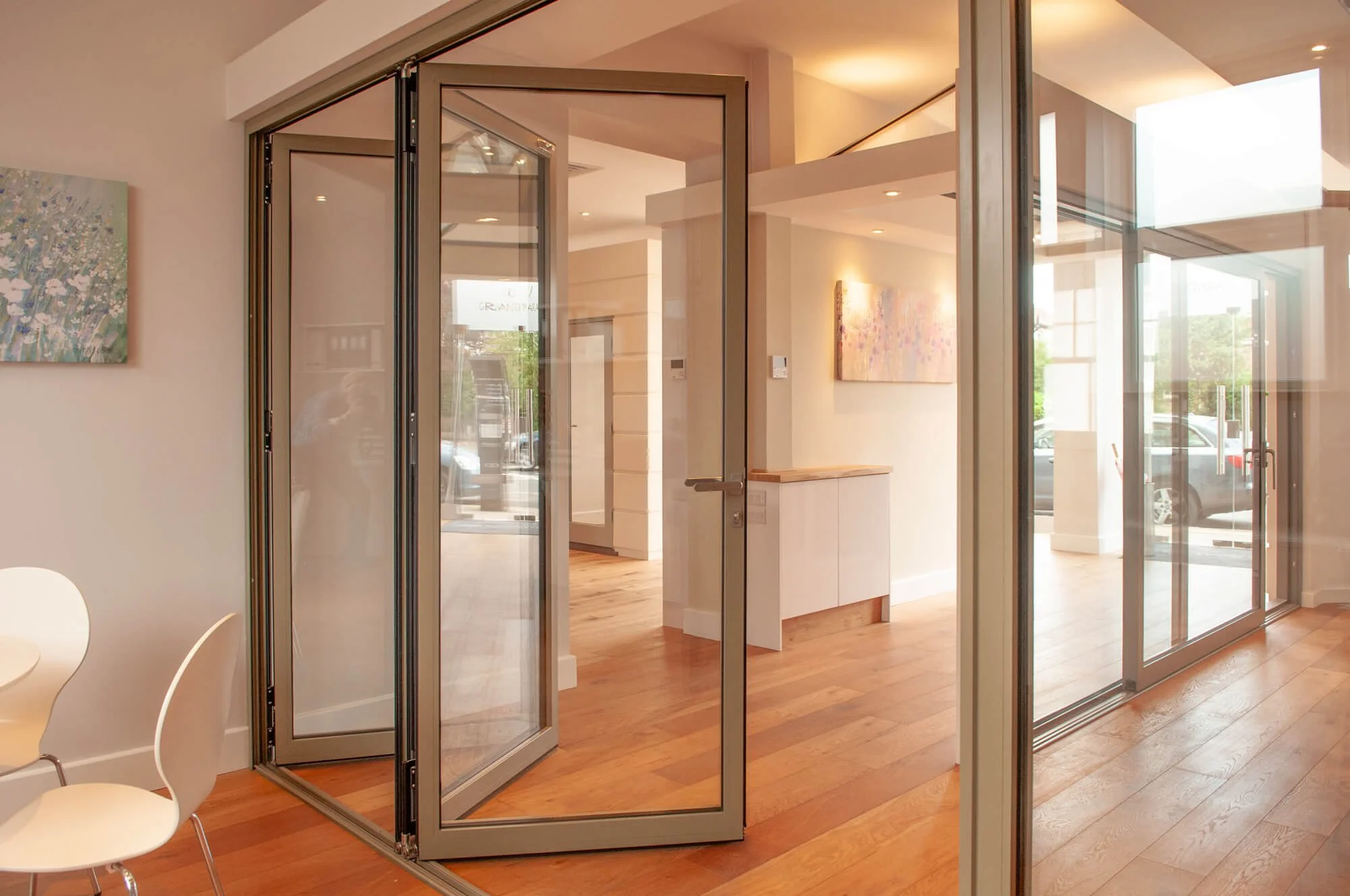 Modern interior space with glass walls and wooden floors, featuring a foldable glass door, chairs, and paintings on the walls.