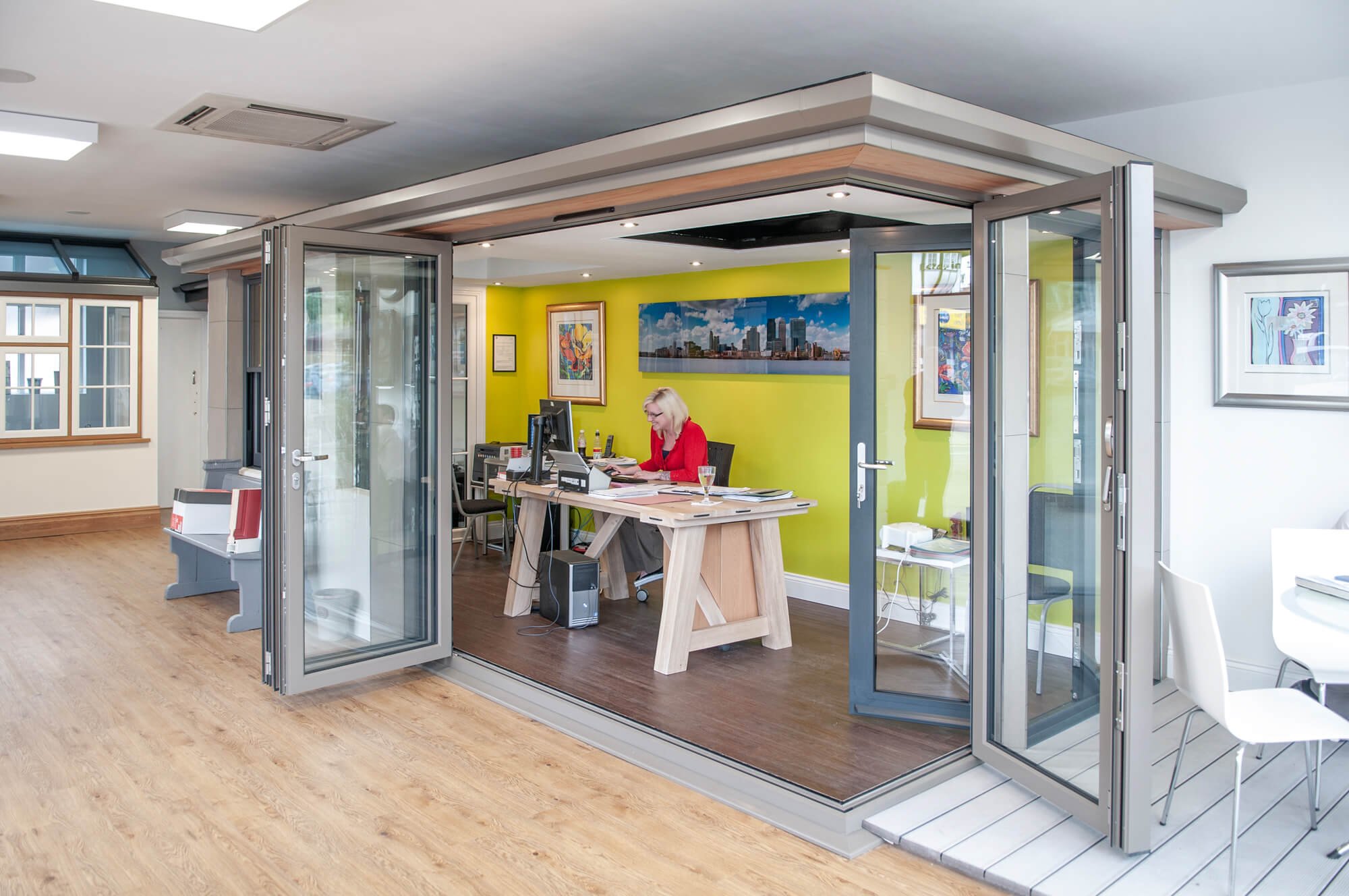Open glass door leading to a modern office with a woman working at a desk with computer, bright green wall, framed artwork, and wooden flooring.
