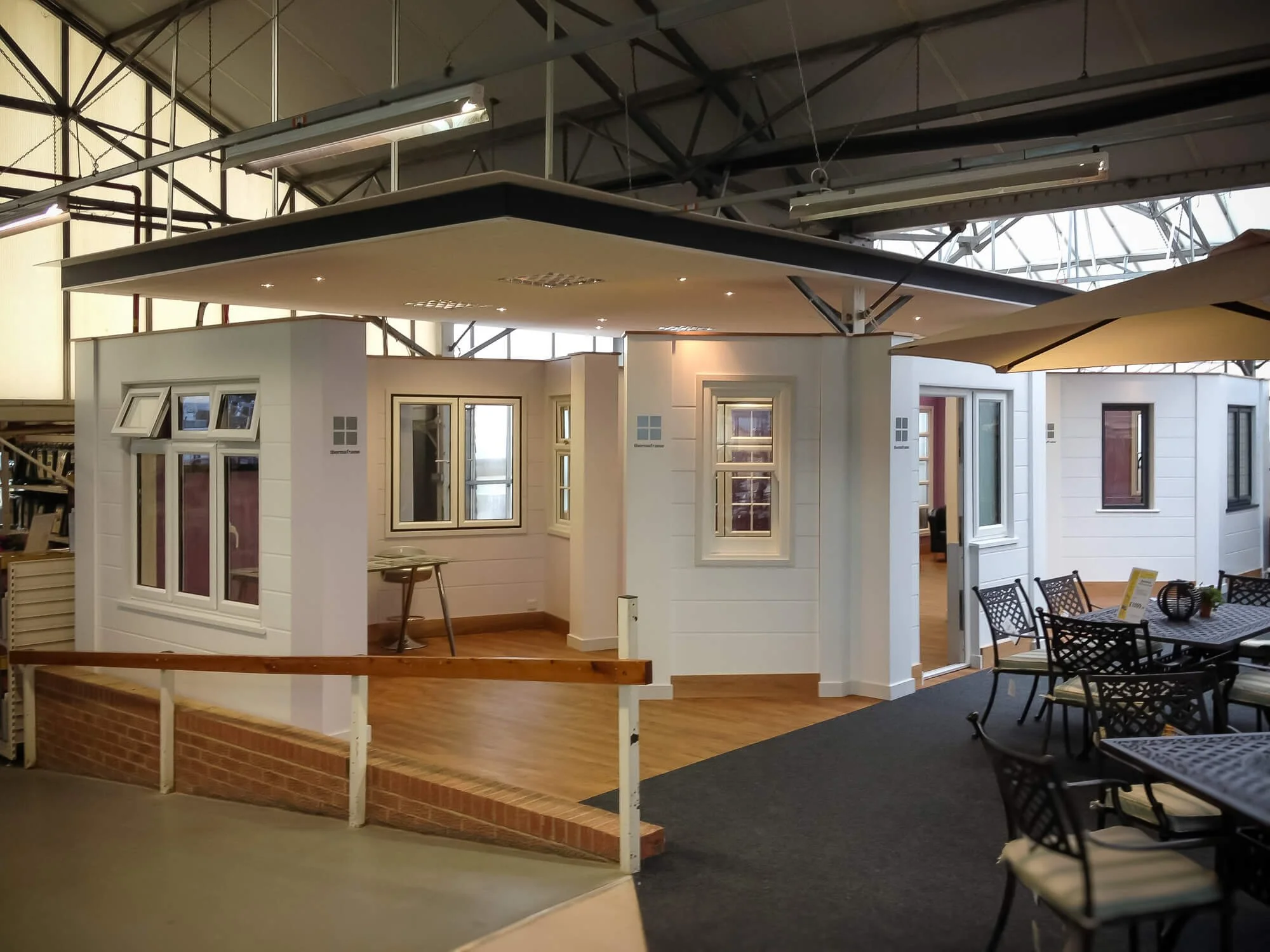 Indoor display of model house with white walls, multiple windows, and outdoor furniture, under a high ceiling with metal framework.