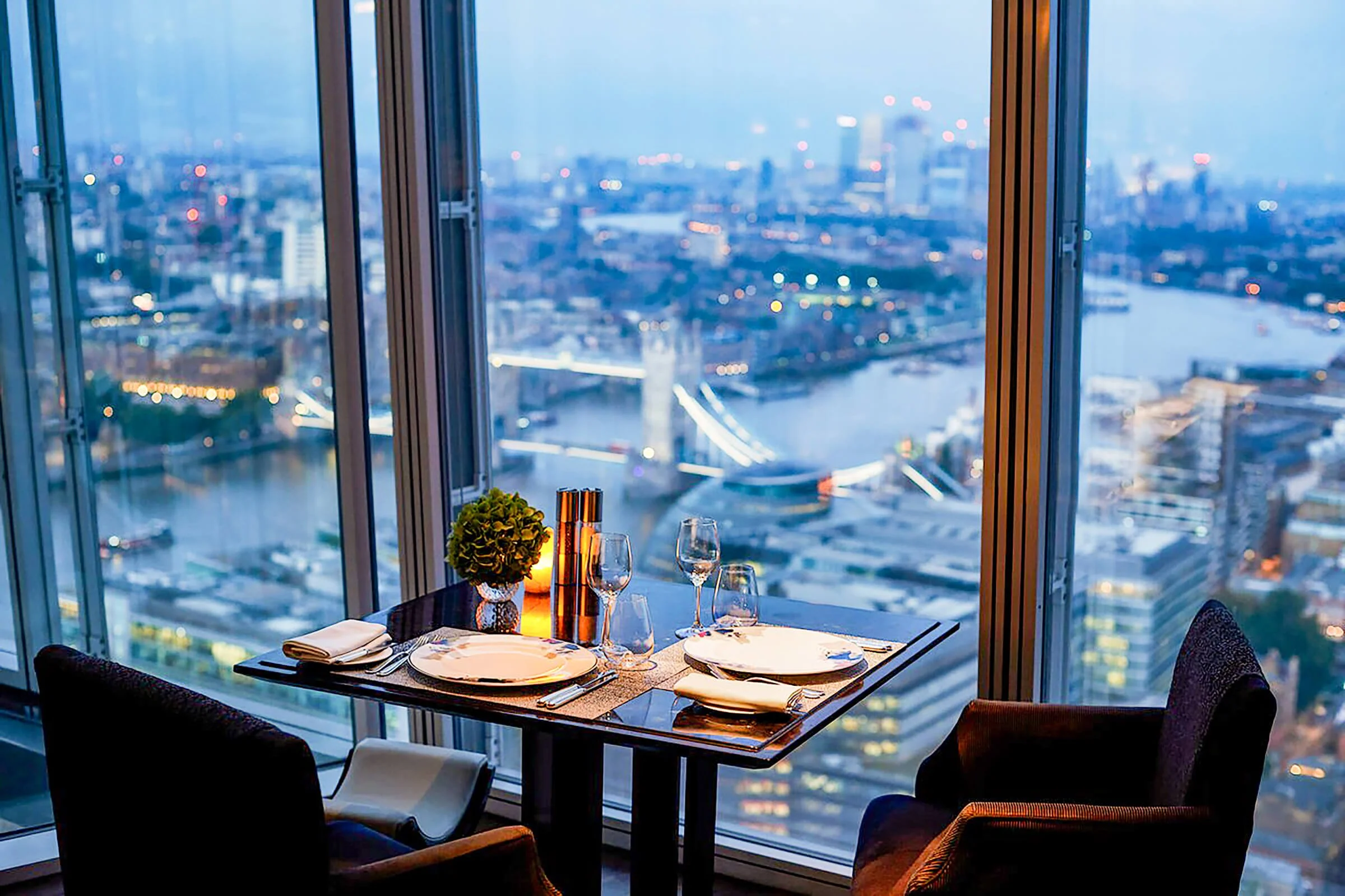 Table set for two with wine glasses, plates, napkins, and a small potted plant, in a high-rise restaurant with a view of a city skyline and river at dusk.
