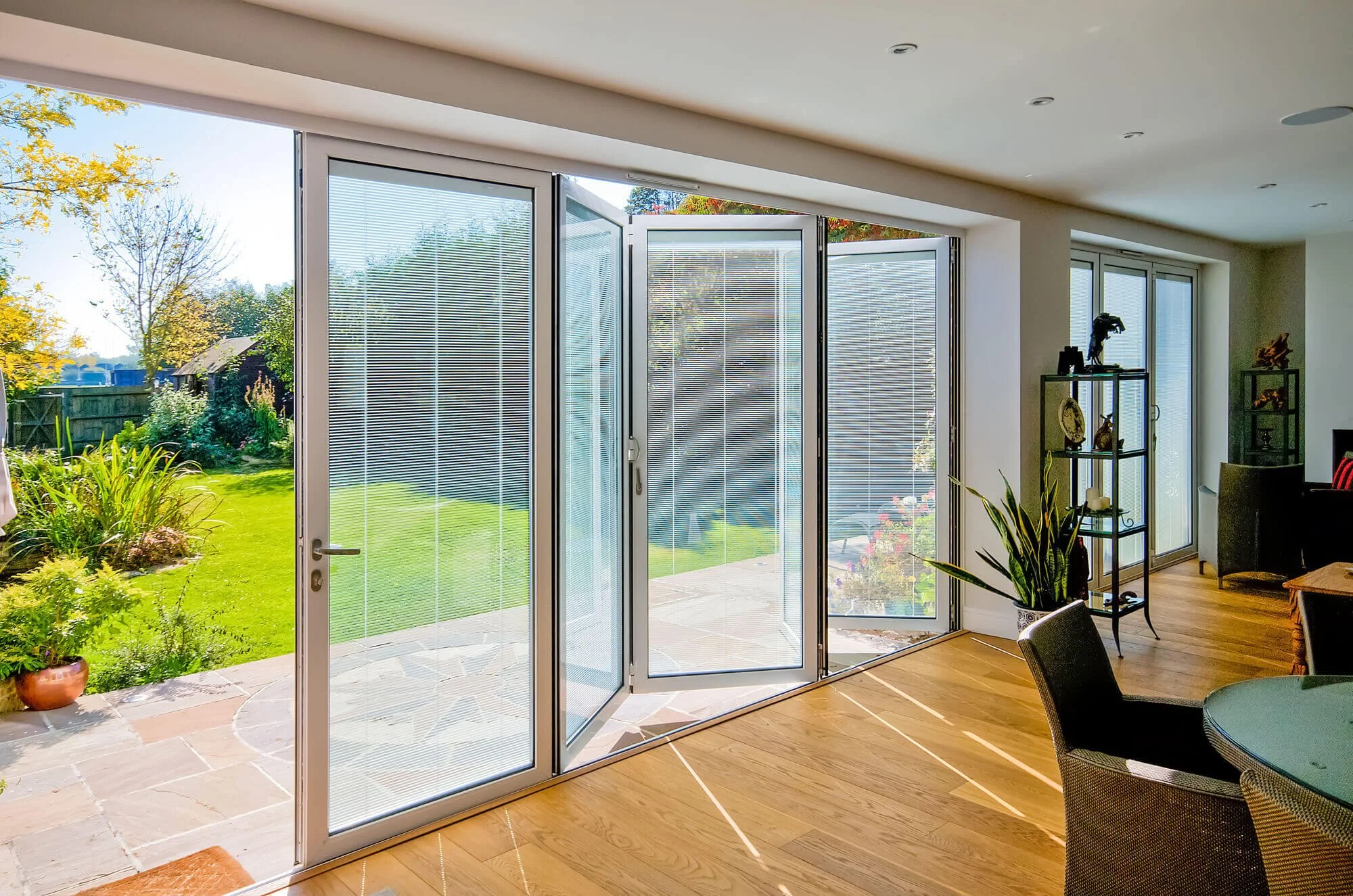 Interior view of a room with large glass doors and windows opening to a green backyard with trees and plants, sunlight casting shadows inside the room, which has wooden flooring and furniture.
