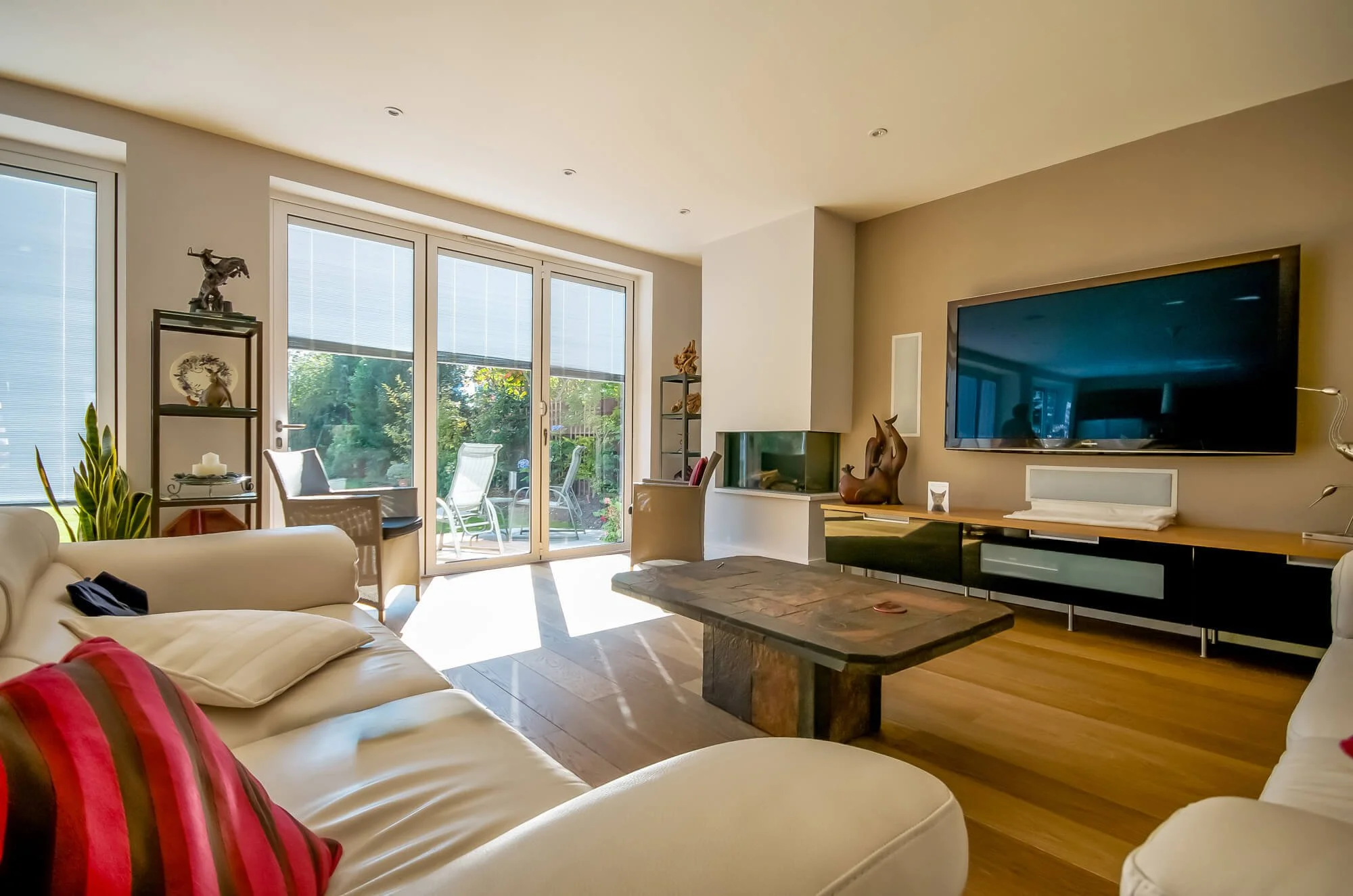 Bright living room with large sliding glass doors, white couches with red and black pillows, central stone coffee table, wall-mounted TV, and patio furniture outside.