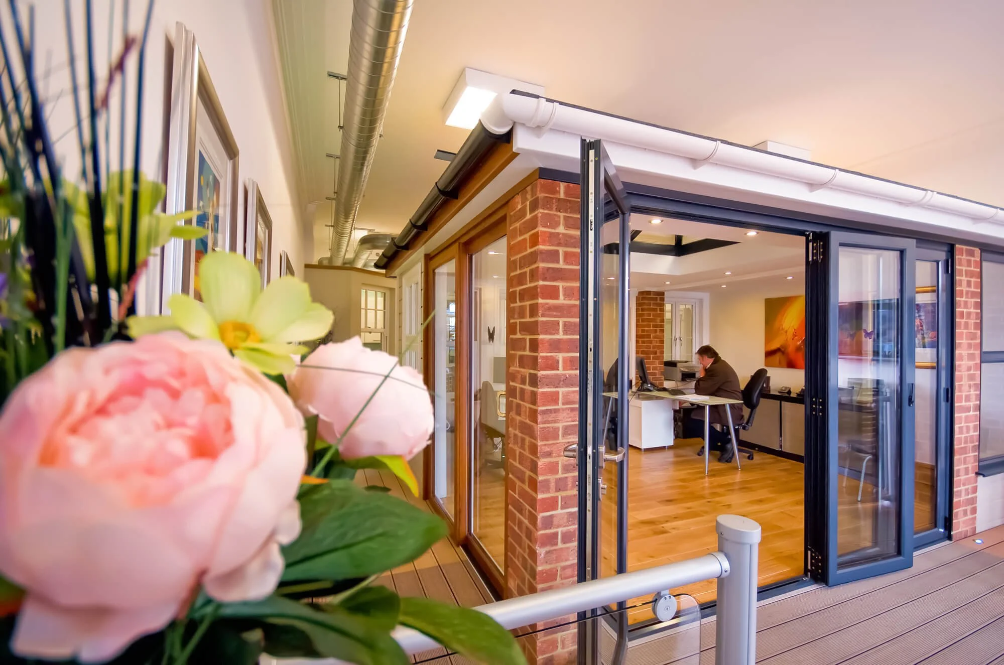 Office space seen through open glass doors with brick and wood walls, a person working at a desk, and a large abstract painting on the wall.