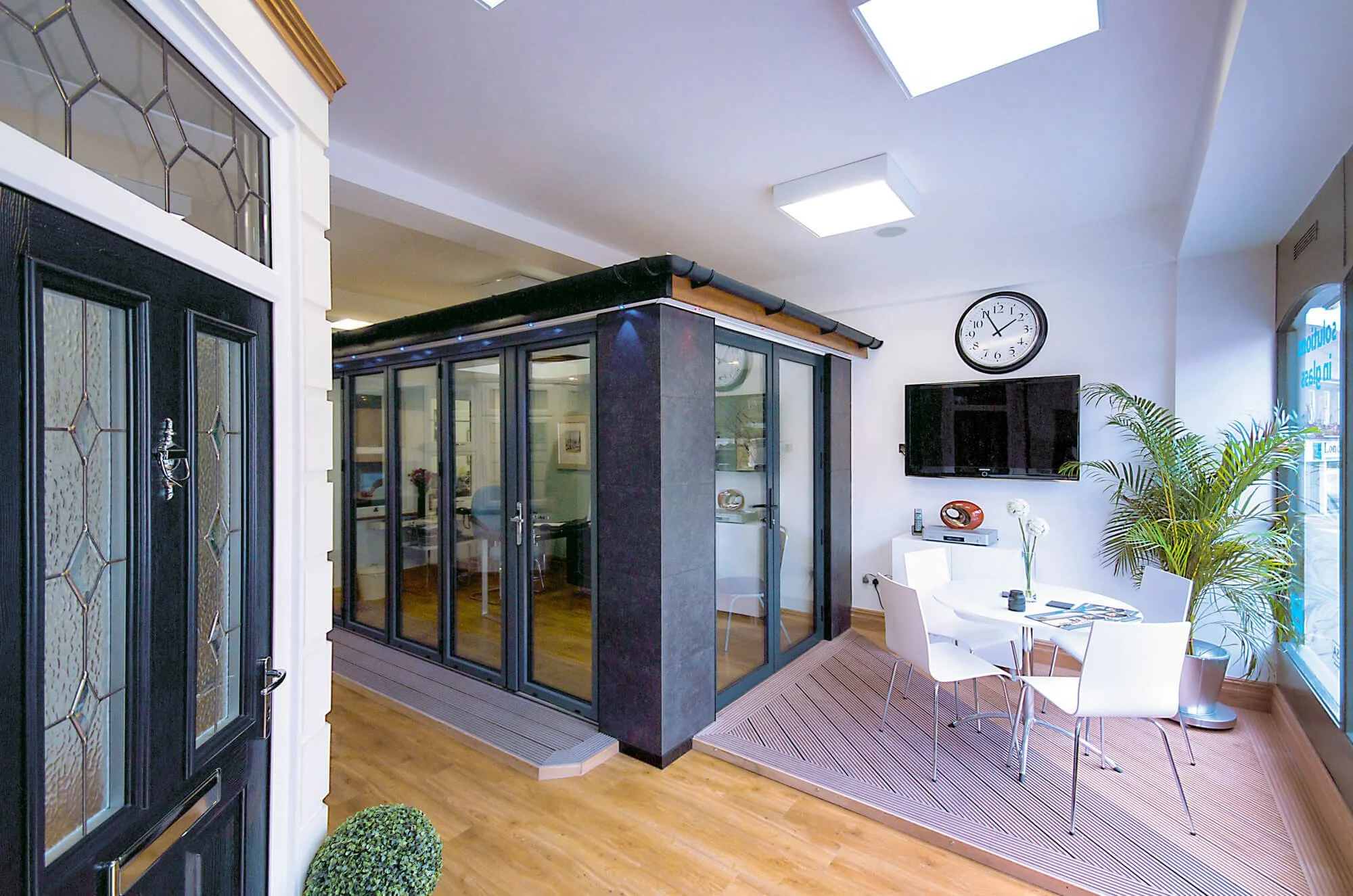 Interior view of a modern office space with a glass-enclosed meeting room, a wall-mounted TV, a round dining table with four chairs, a large potted plant, and window for natural light.