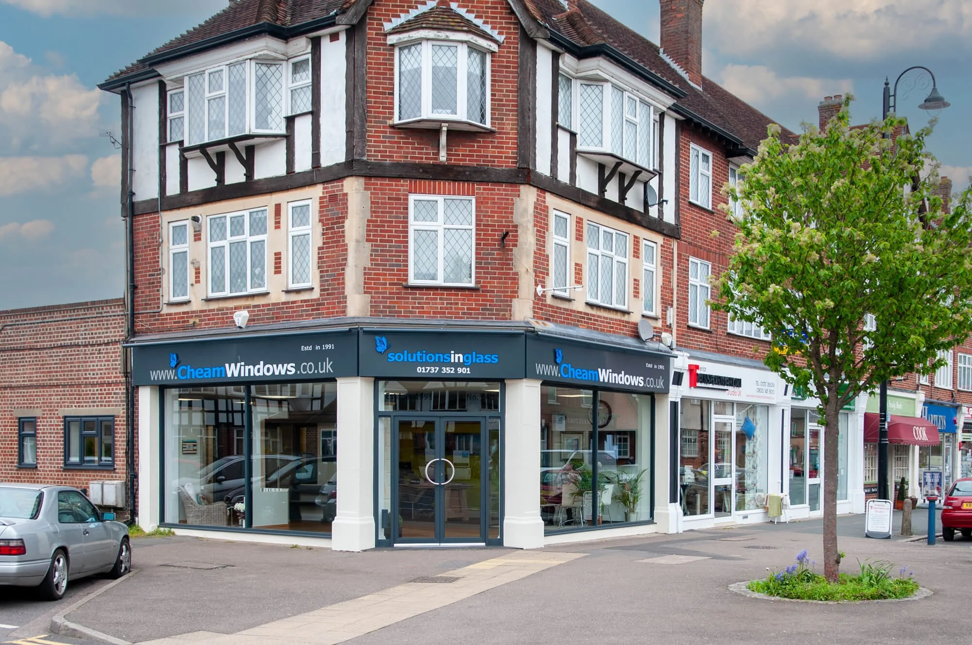 Street scene with a corner building housing a window business named Cheam Windows, with red brick and black timber accents, parked cars, a small tree, and several other storefronts.