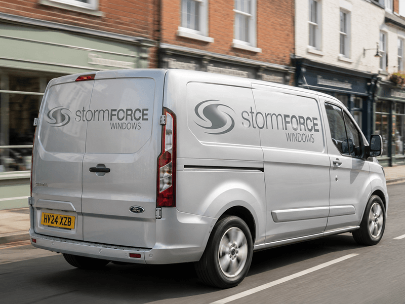 A white Ford Transit van driving down a city street, with the logo and text for Stormforce Windows on the side and back.
