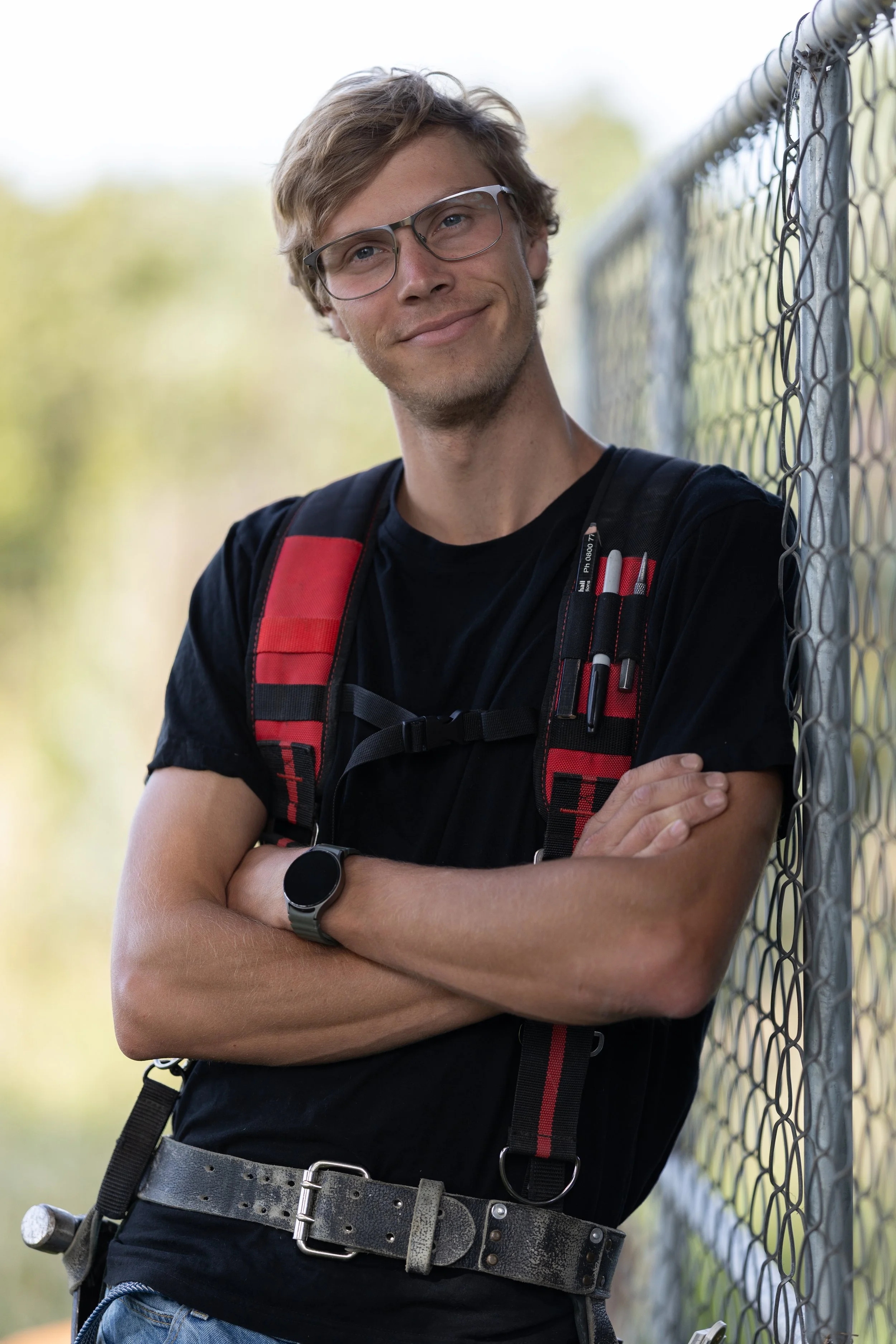 A young man with blond hair, glasses, and a slight smile, standing outdoors next to a chain-link fence. He is wearing a black T-shirt, a red and black backpack, a black watch, and a black belt with a silver buckle, with one arm crossed over the other.