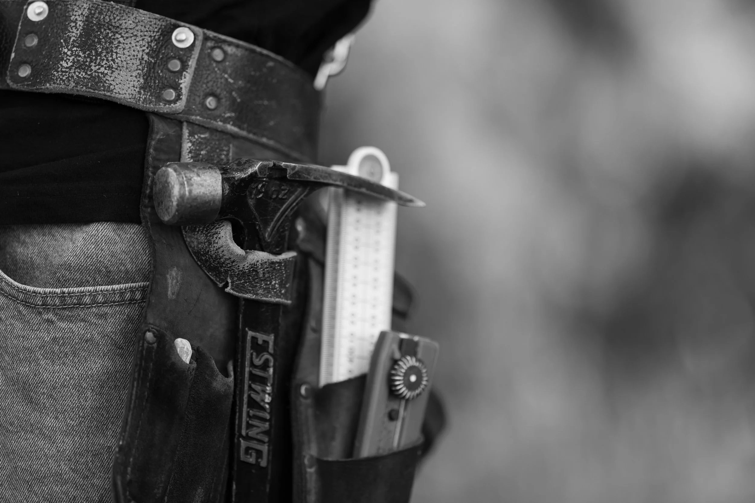Close-up of a person's waist and hip area, showing a worn black leather tool belt with metal rivets, a hammer, and a measuring tape attached, wearing denim jeans.