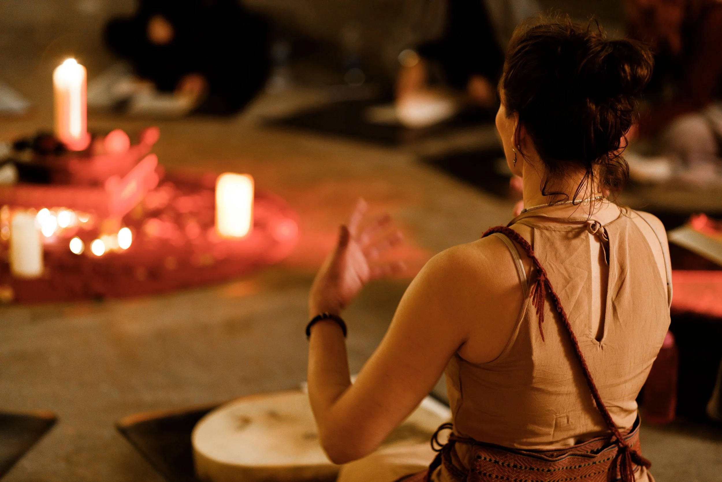 A woman is standing with her back to the camera in a dimly lit room with candles and blurred figures in the background, appearing to be engaged in a spiritual or meditative practice.