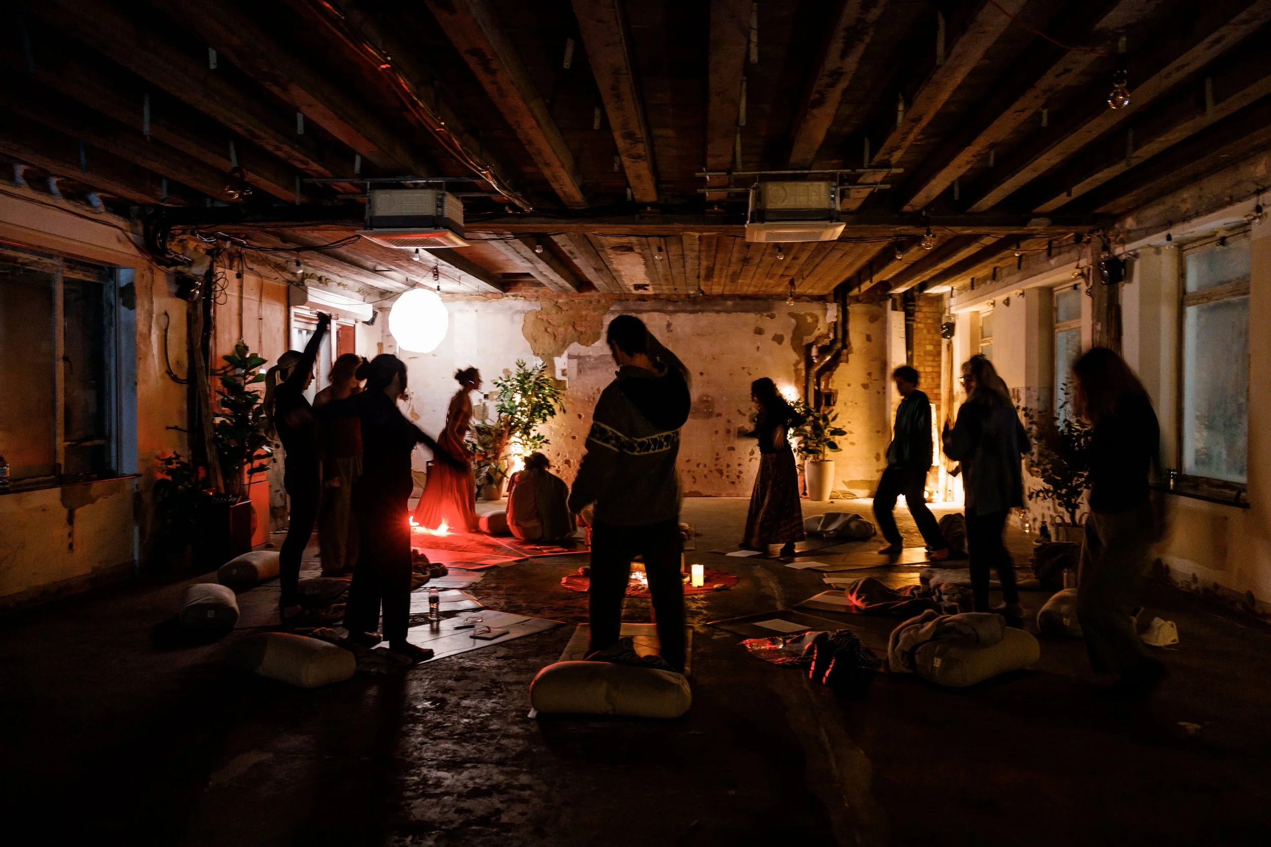 People are gathered in a dimly lit indoor space with exposed brick and wooden ceiling beams. They appear to be participating in a spiritual or meditation session, with some sitting on cushions and others standing. There are plants, candles, and hanging lights creating a warm, relaxed atmosphere.