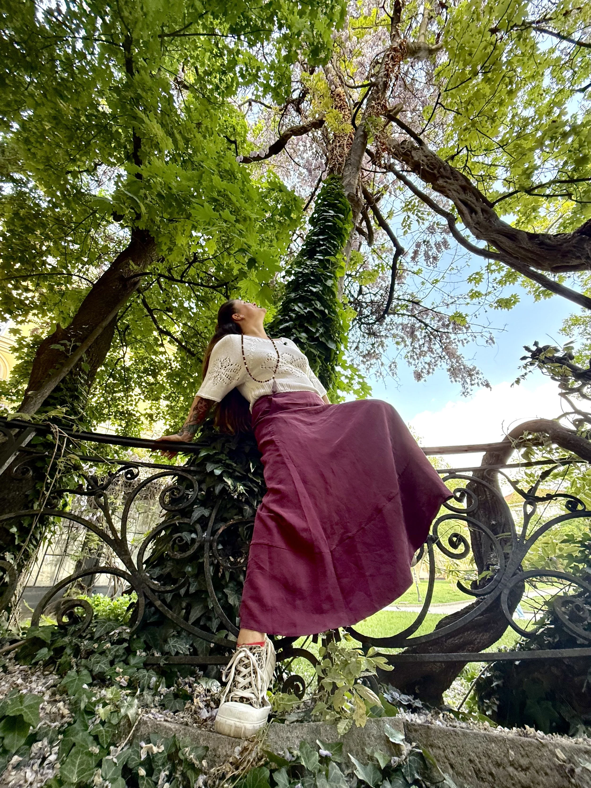Woman sitting on a decorative wrought iron fence beneath leafy green trees, wearing a white blouse and a long maroon skirt, with one foot on the ground and one foot resting on the fence, looking upwards.