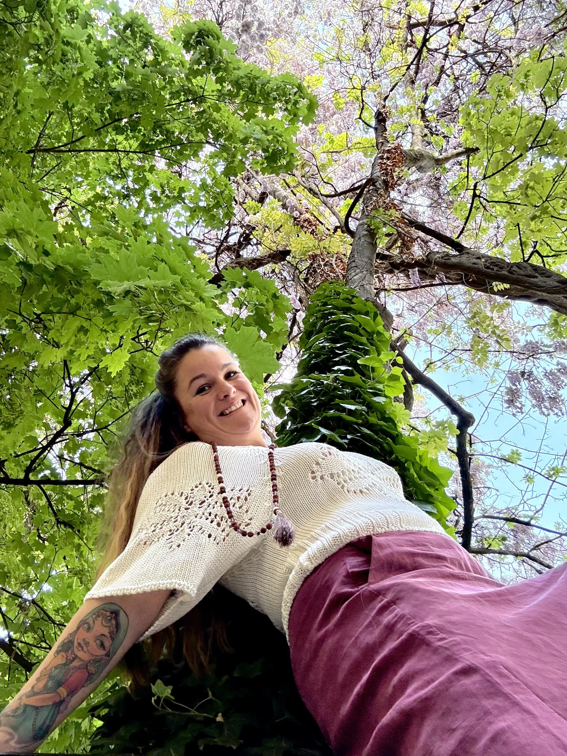 A woman with long hair, smiling, wearing a white sweater with lace details, maroon pants, and a beaded necklace, looking down at the camera from below in an outdoor setting with green trees and sky in the background.
