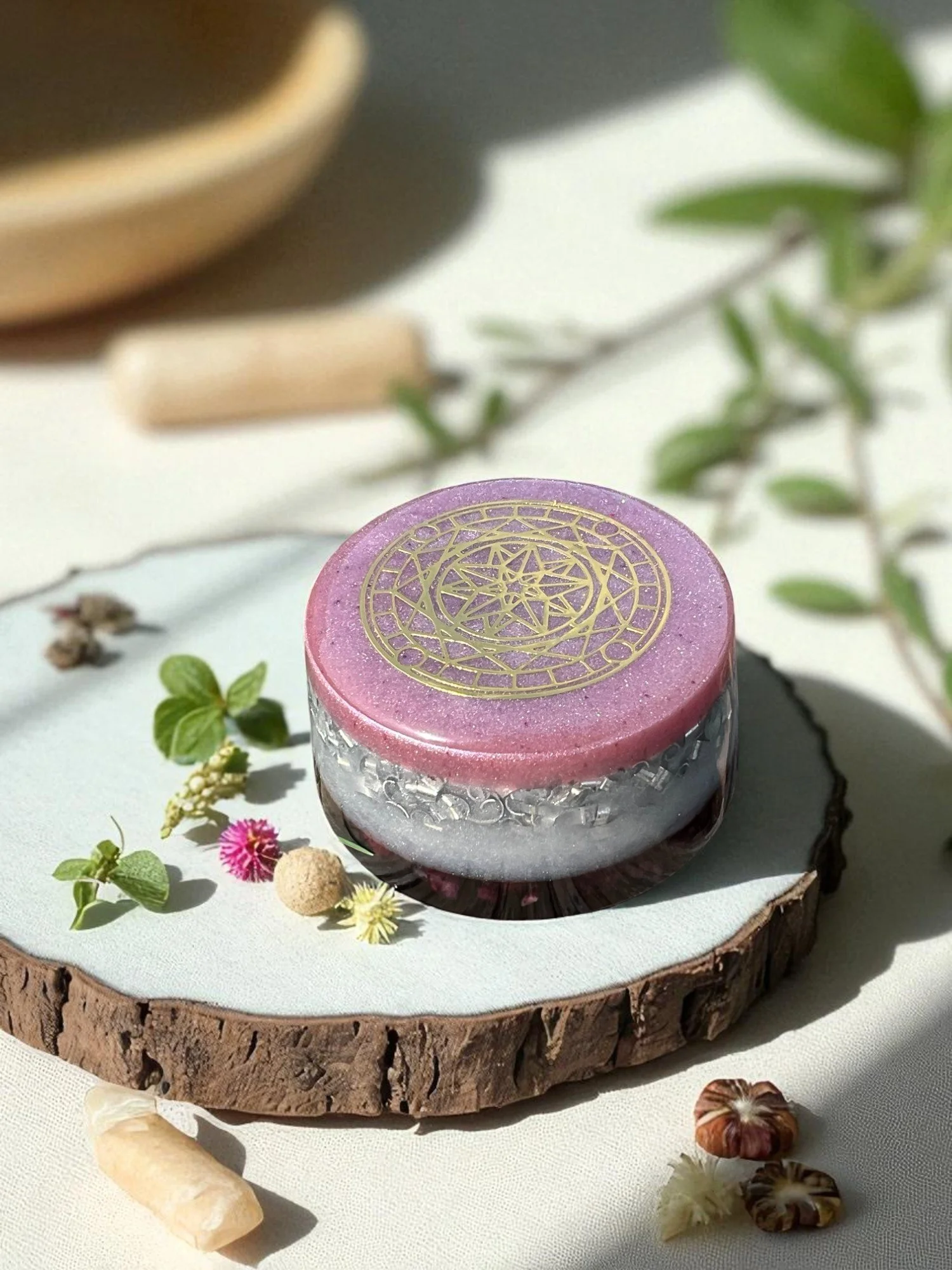 A pink and silver cylindrical candle with a gold geometric design on top, placed on a wooden slice with plants and small stones nearby.