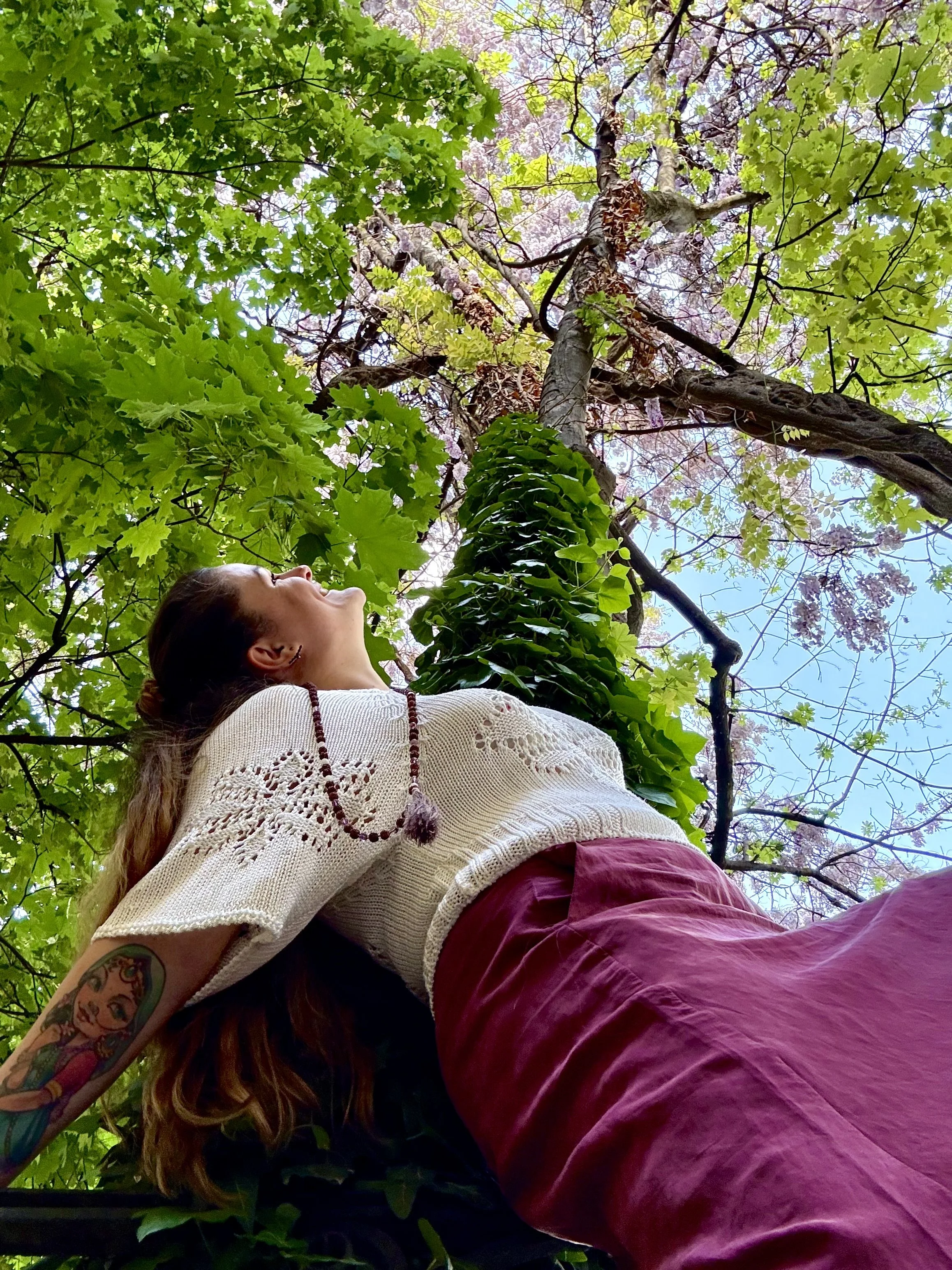 A woman with long brown hair and tattoos on her arm is lying on her side under leafy tree branches, looking up at the sky, wearing a white crocheted top and burgundy pants.