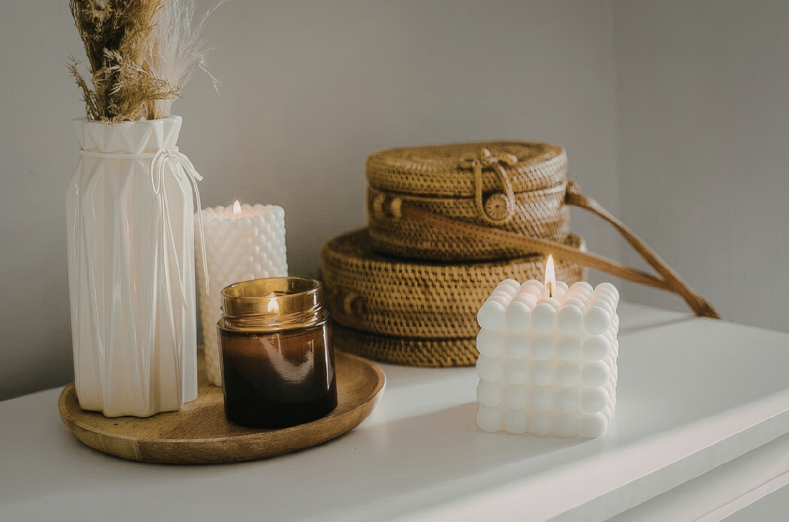 Decorative arrangement on a white surface featuring a tall white vase with dried grasses, a white textured candle with a flame, a small brown candle jar on a round wooden tray, and two woven baskets with lids, one stacked atop the other, with a white bubble cube candle with a flame in front.