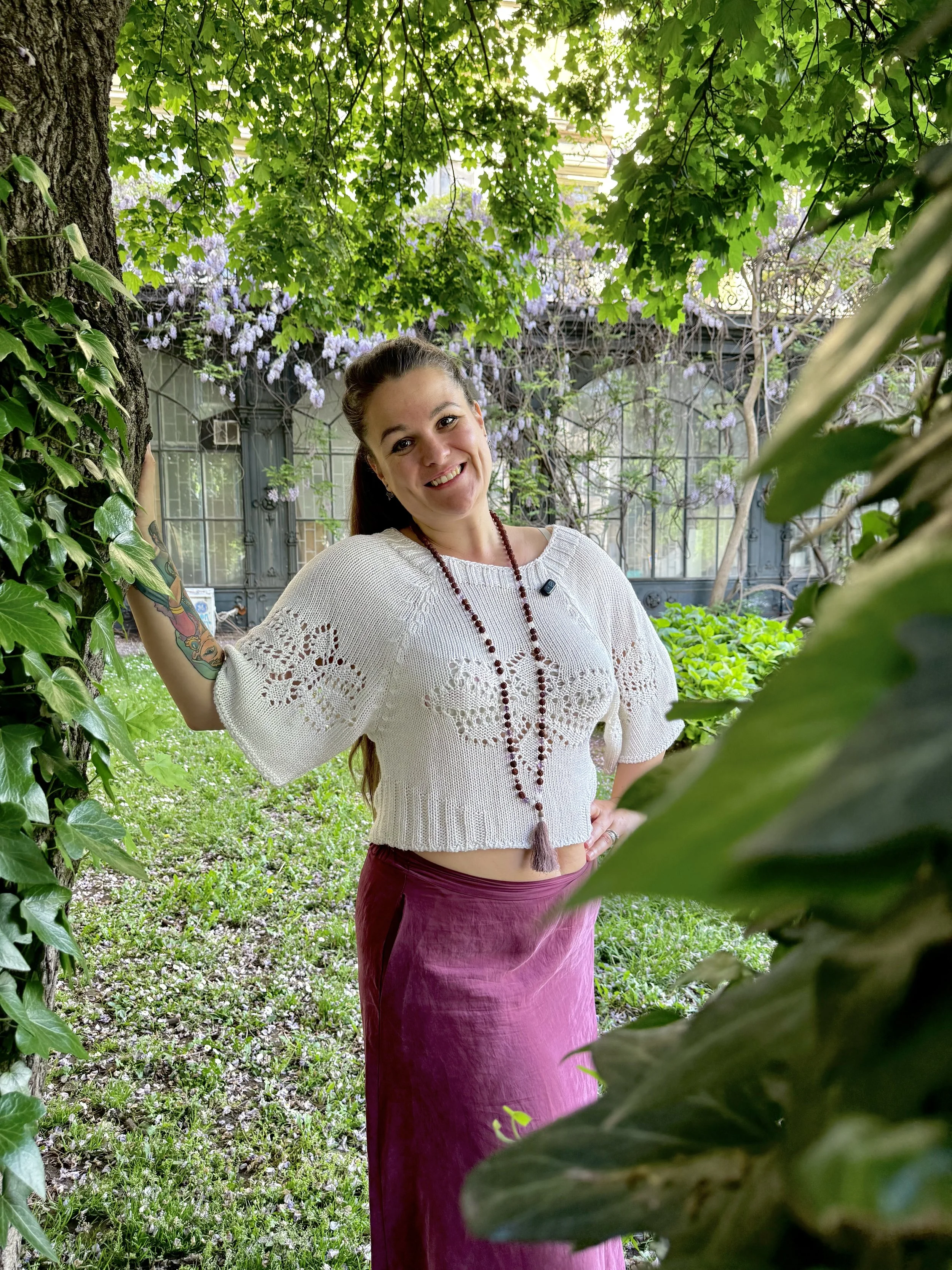 A woman with brown hair, tattoos on her arm, wearing a white knit top and a long pink skirt, smiling and leaning against a tree with green leaves and purple flowers in a garden setting.