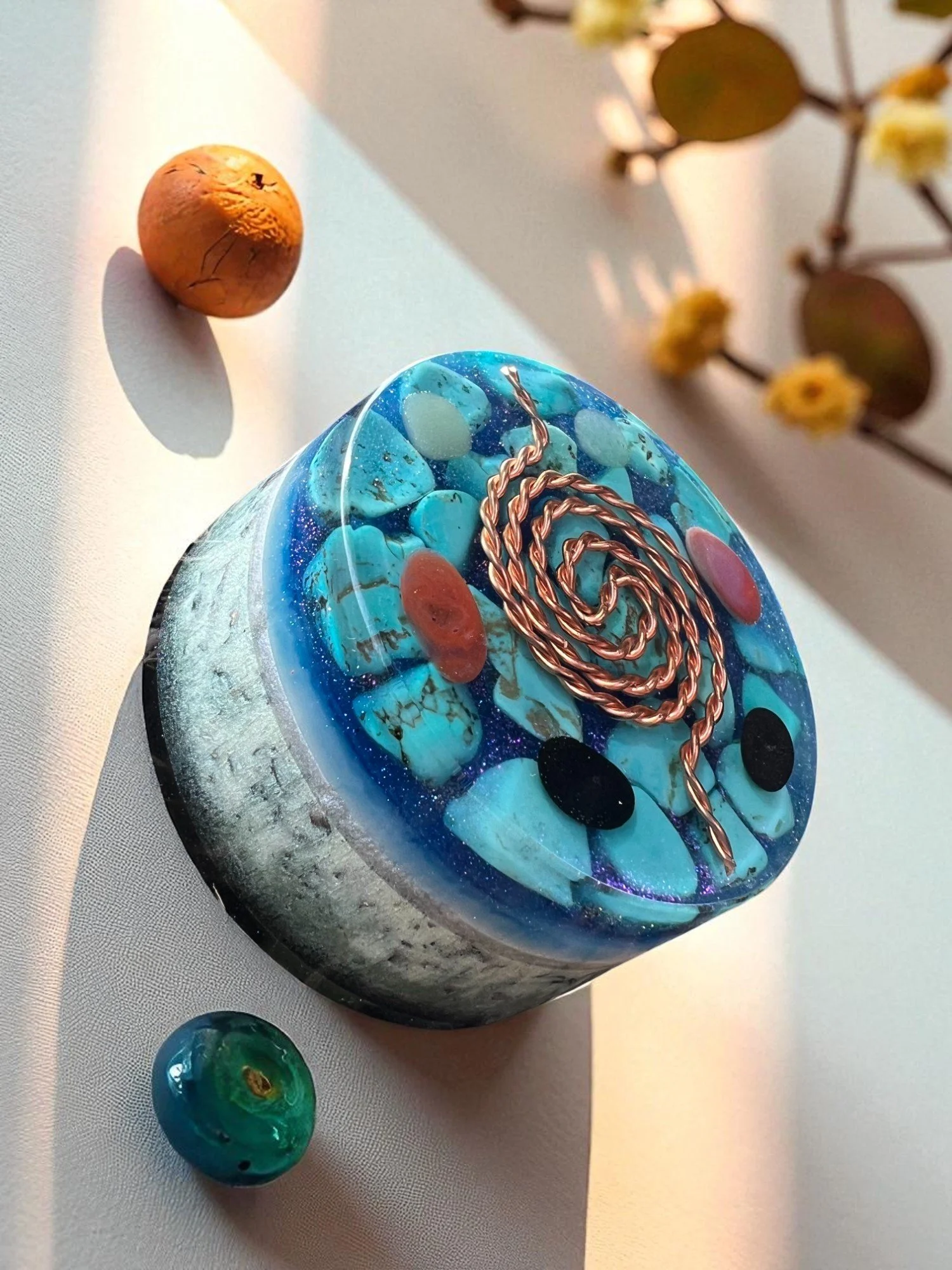 An artistic glass sculpture with blue, turquoise, and pink colors, featuring copper wire details. A small pebble and a decorative glass marble are also on a white surface, with a blurred floral arrangement in the background.