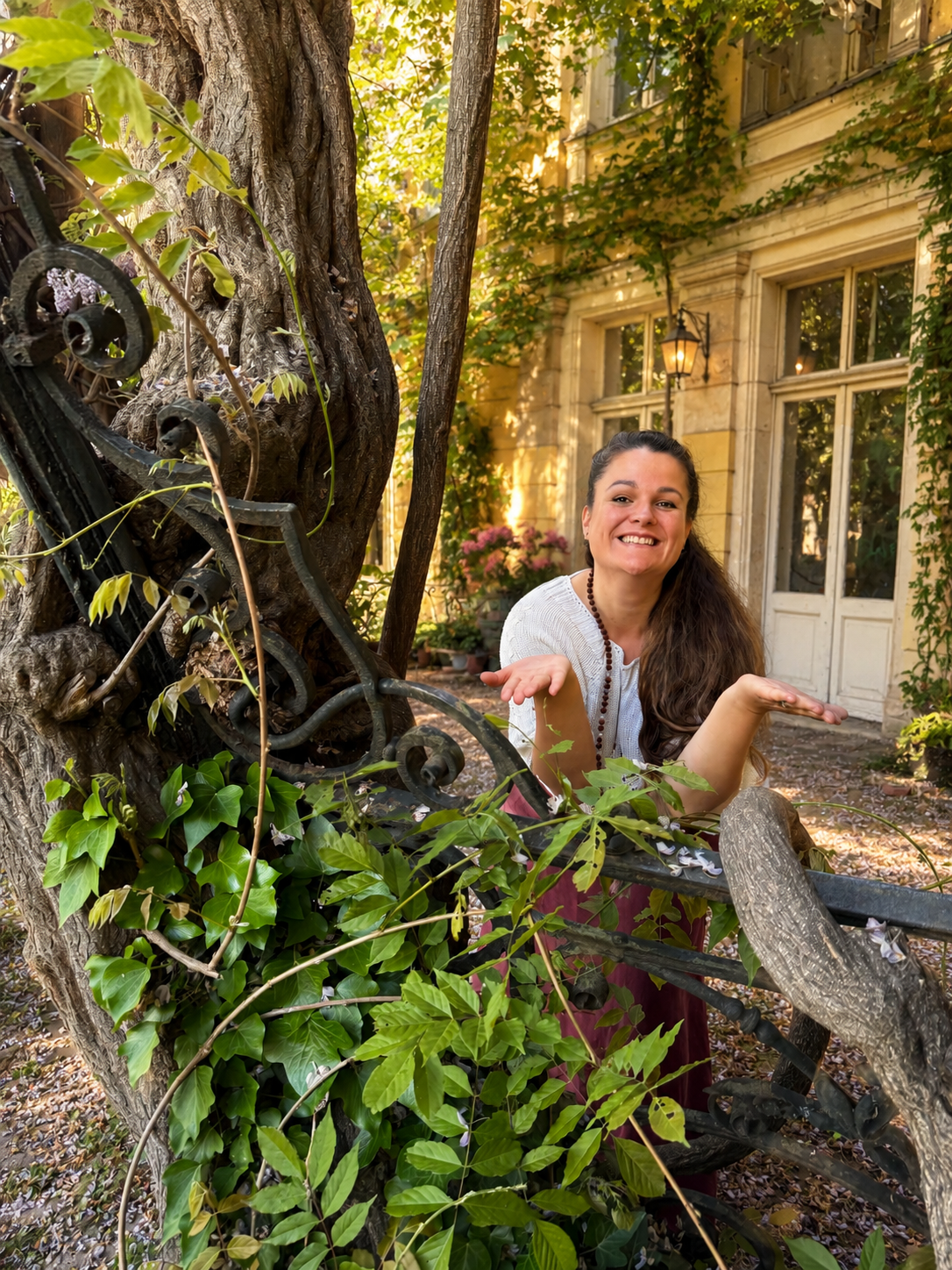 A smiling woman with long brown hair, wearing a white top and maroon skirt, stands outdoors near a large tree with ivy, in front of an old building with large windows and ivy growing on the walls, during fall.