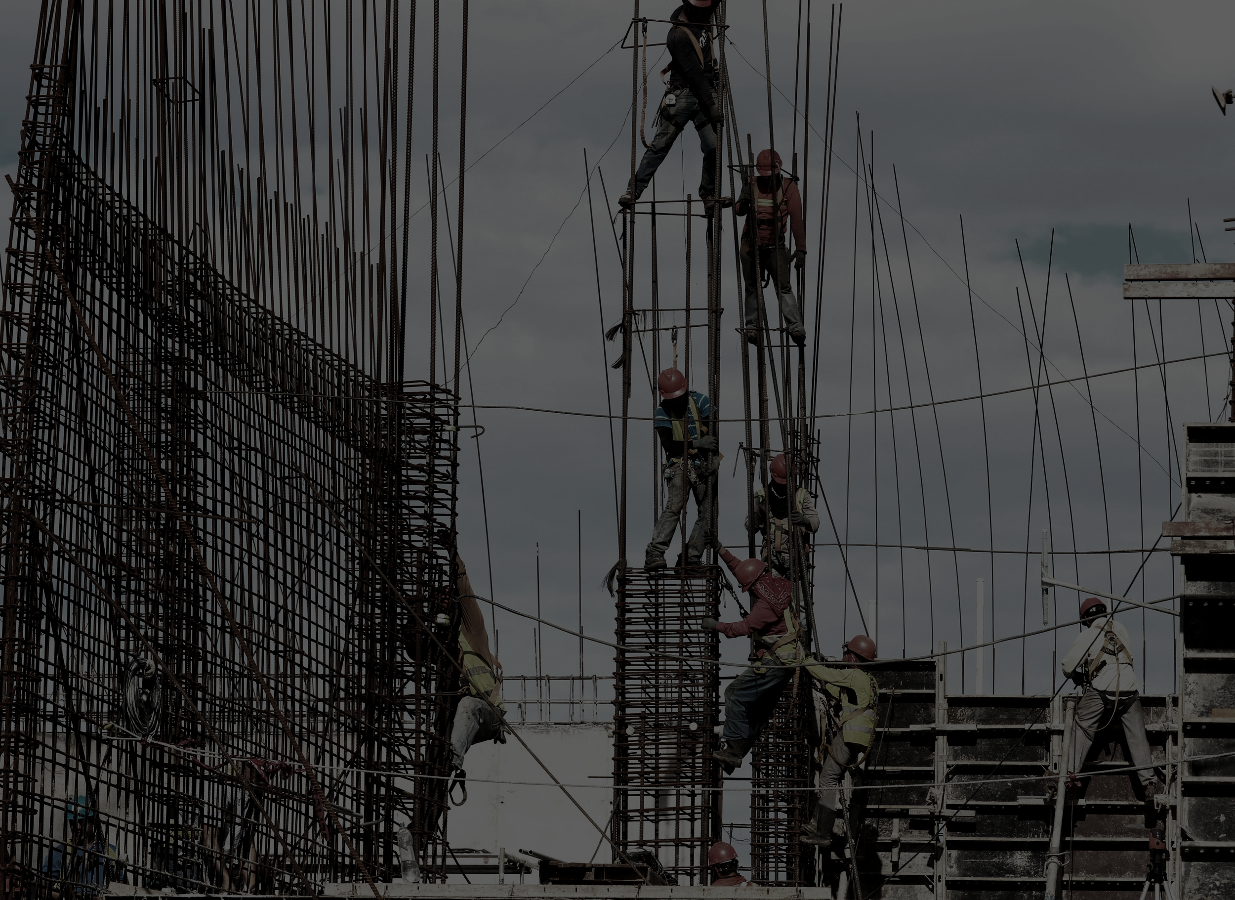 Construction workers wearing safety gear are assembling steel reinforcement bars on a building site under cloudy skies.