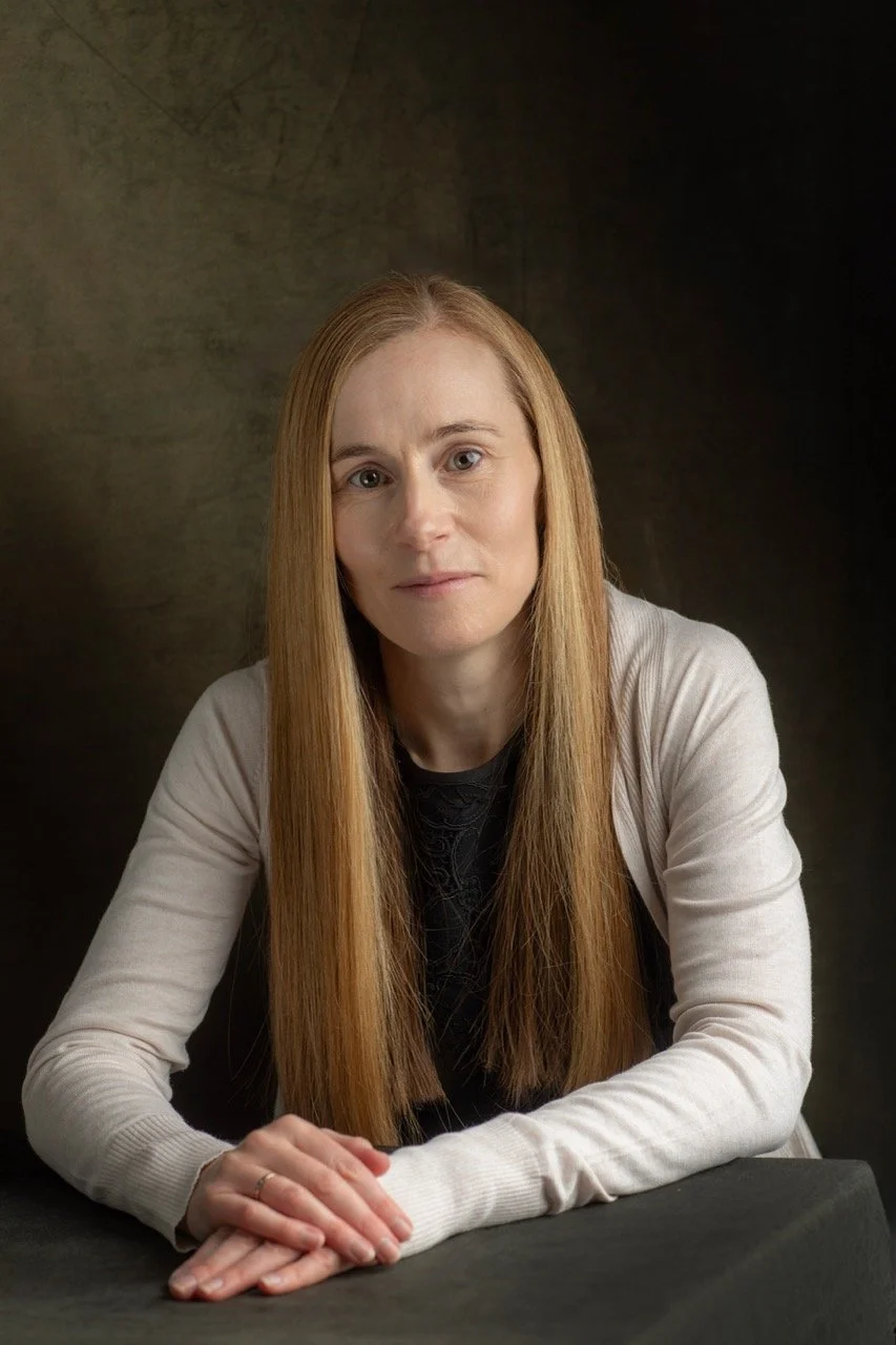 Female mental health nurse with long blonde hair looks at the camera against a mottled brown backdrop. She is wearing a black t-shirt and cream cardigan.