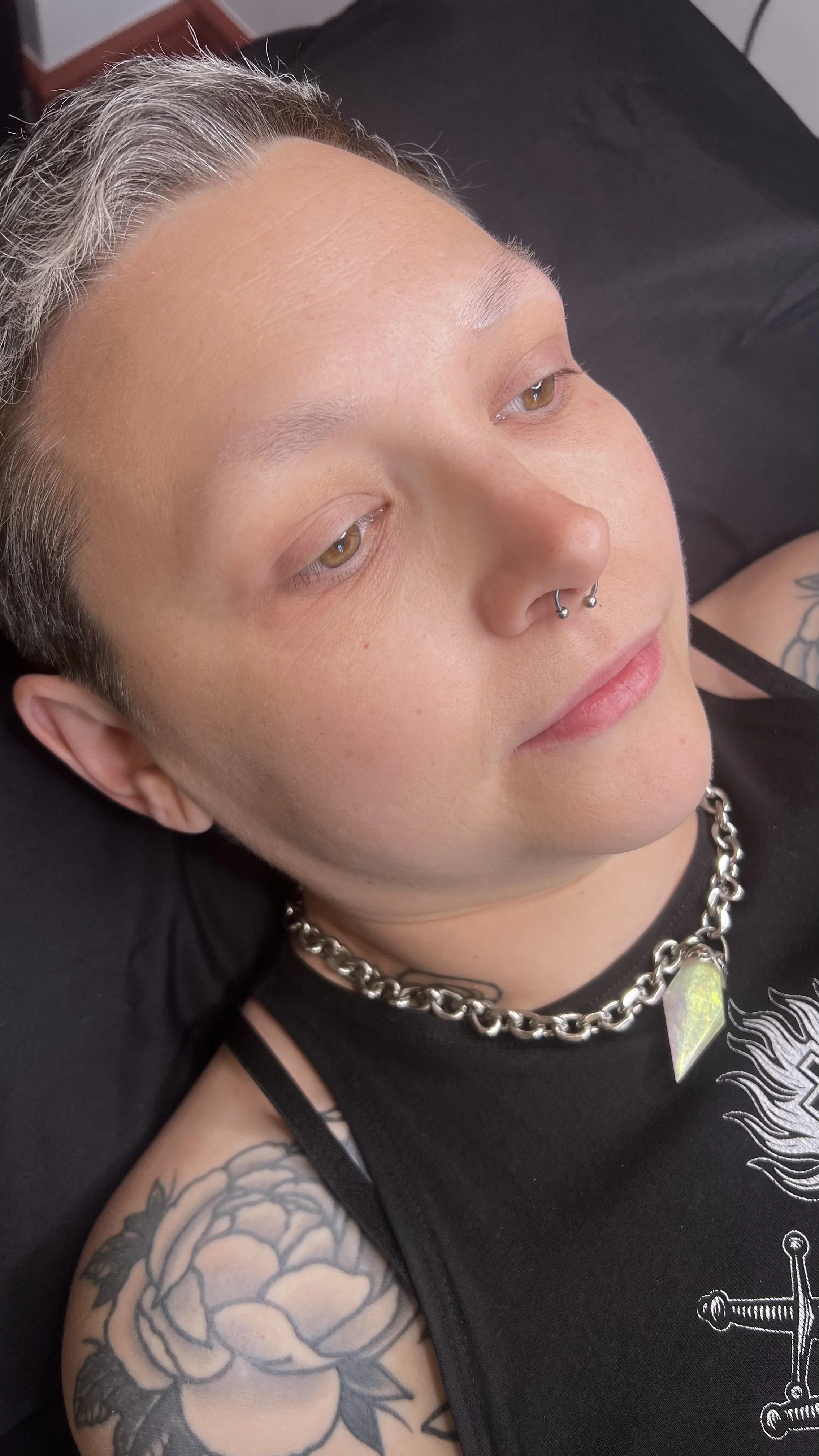 A person with short gray hair and light skin lying down with closed eyes. They have a septum piercing, a silver chain necklace with a large iridescent pendant, and a black sleeveless shirt with a graphic design. They have floral tattoos on their upper arm.